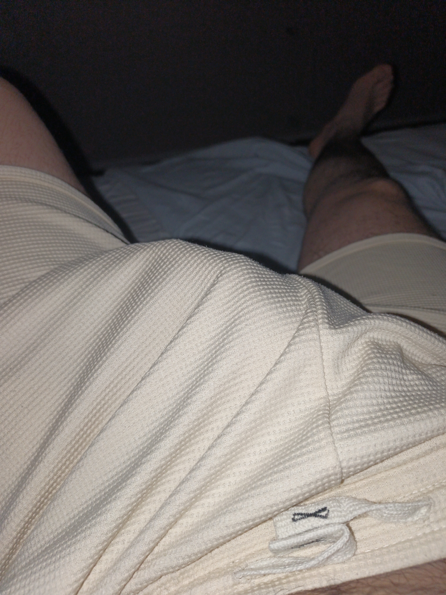 The image shows a close-up view of a person's legs and upper body wearing light-colored, textured shorts. The person is lying down on a bed with blue sheets. The background is dark and out of focus. There is a label with an X-shaped symbol on the shorts. The image does not contain any information about the location in Ümraniye, Turkey.