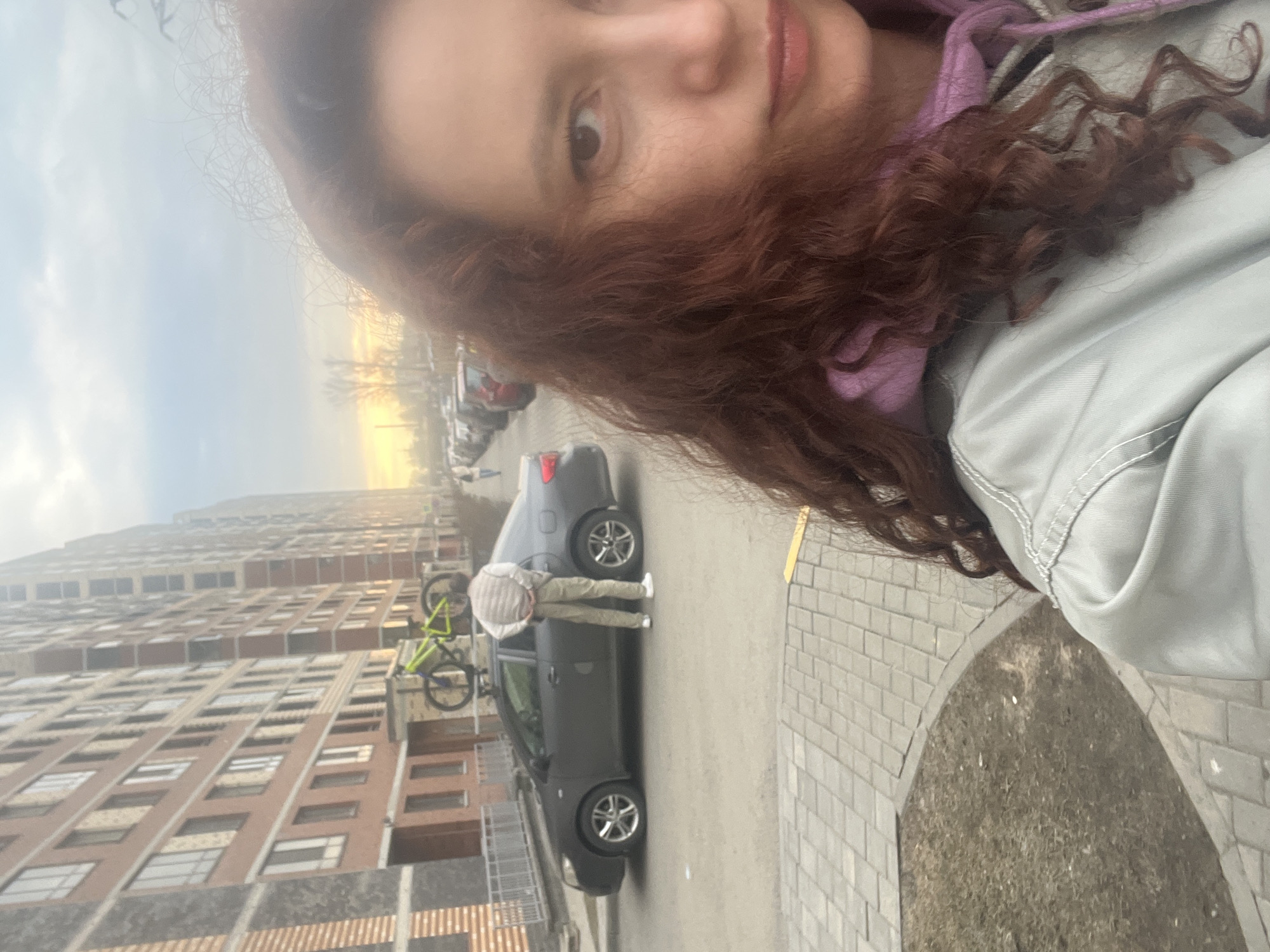 The image depicts a woman with curly brown hair in an outdoor setting. She appears to be taking a selfie, as her face is prominent in the foreground. Behind her, a gray car is parked on a paved area next to a tall apartment building. A person is bent over near the car, and a green bicycle is mounted on the vehicle. The scene is set in Saint Petersburg, Russia, evidenced by the architecture of the apartment building and the streetscape. The weather appears overcast with a hint of sunset visible in the sky.