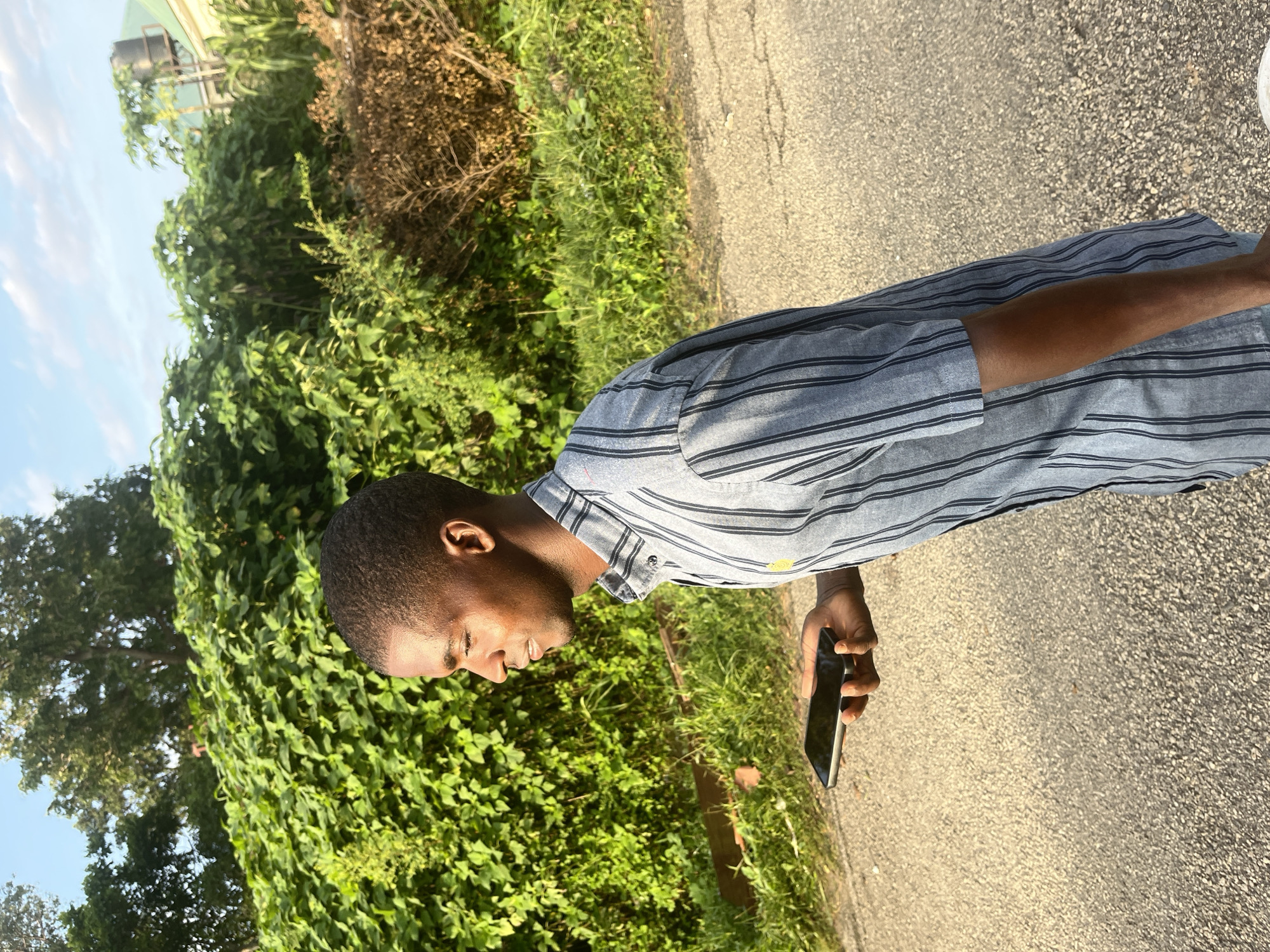 The image depicts a young Black man walking on an asphalt road, holding a black smartphone in his left hand. He is wearing a short-sleeved, light blue striped shirt with darker blue vertical stripes and a small yellow emblem on the chest. The man is in profile, looking to his left. Behind him, a dense green bush and trees form the backdrop, with some dry brown foliage also visible. In the distance, part of a building with a green roof and a water tank can be seen, suggesting a semi-urban or suburban environment. The lighting suggests it is late afternoon, with the sun casting a warm glow on the scene. The location is identified as Abuja, Nigeria.