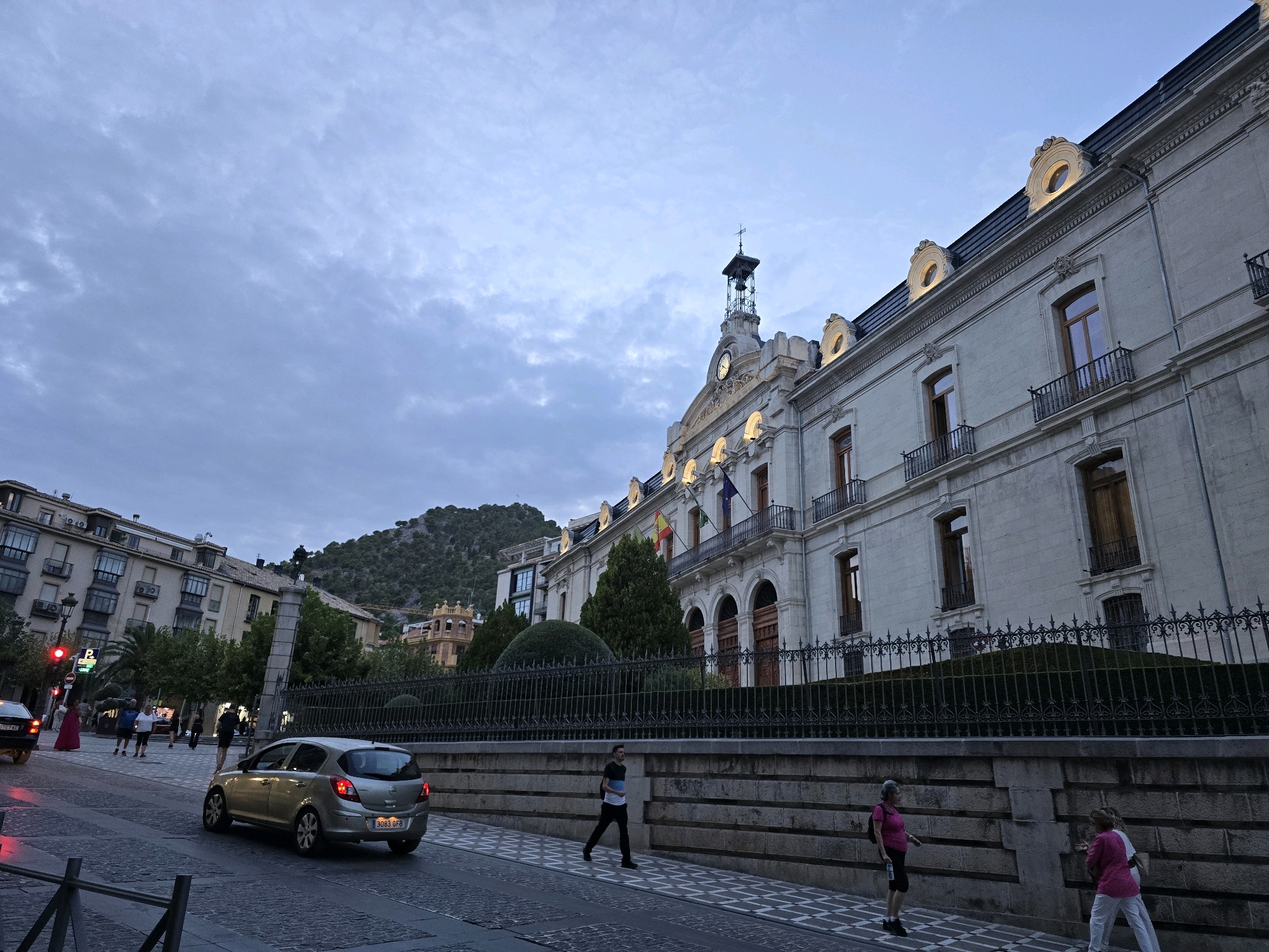 The image shows a street scene in Jaén, Spain, dominated by the Palacio de la Diputación Provincial de Jaén. The building, a grand neoclassical structure, occupies the right half of the frame. It features a light-colored facade, arched ground-floor entrances, multiple windows with balconies, and a prominent clock tower topped with a cross. The sky above is overcast with scattered clouds.
In the foreground, a silver hatchback car is driving away from the camera on a cobblestone street. The sidewalk to the right of the car is paved with white and gray checkered tiles. Several people are walking on the sidewalk. A man wearing a backpack and dark clothing is walking towards the right. Further down, two women in bright pink tops are walking towards the right.
On the left side of the street, buildings with balconies are visible, along with streetlights and traffic signals. In the background, a green, tree-covered hill rises, and on its slopes, other buildings and structures are visible, including a distinctive yellow tower. The overall scene depicts a city street at dusk or during twilight, with pedestrian and vehicle activity.