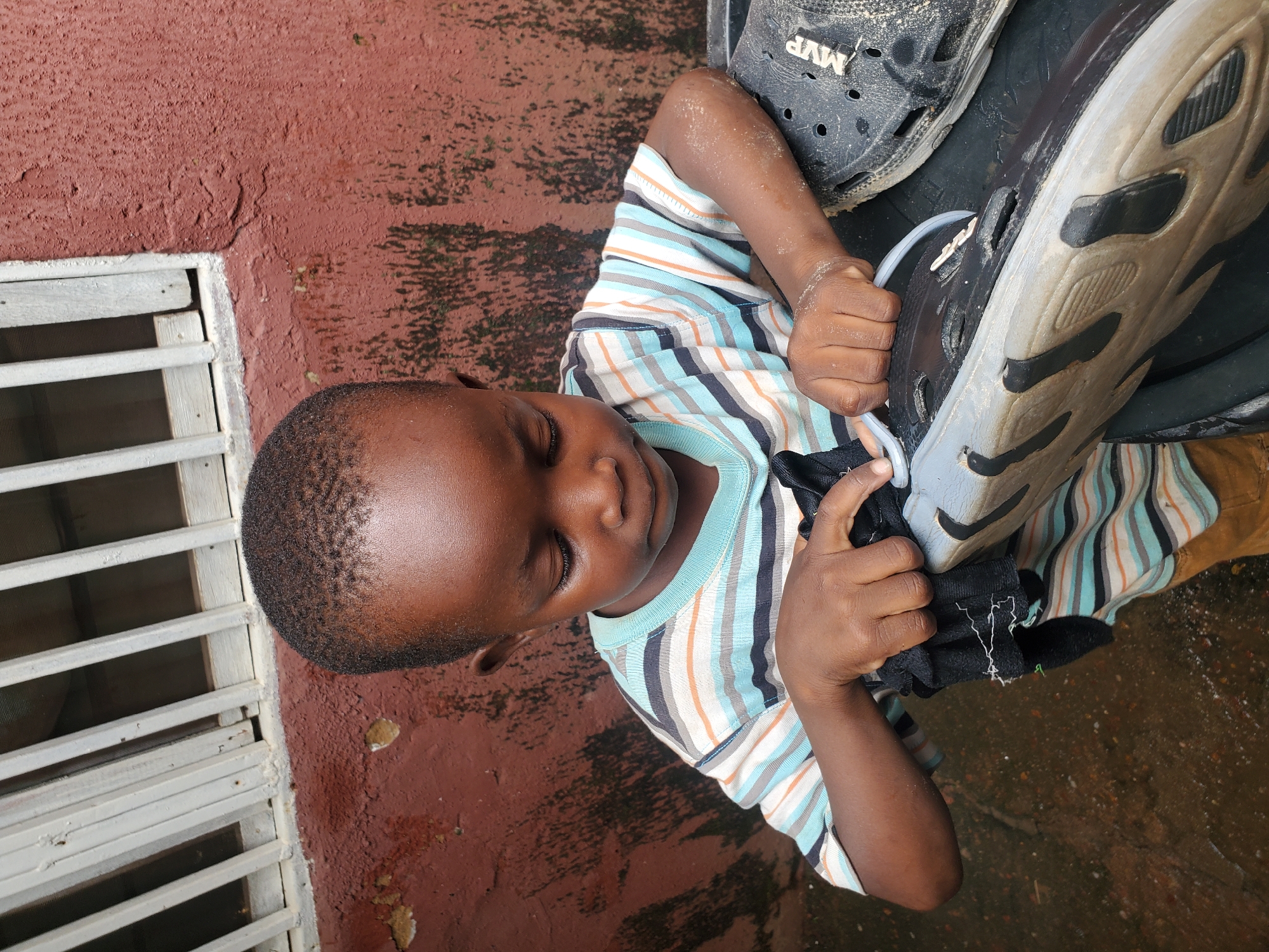 A young boy lies on a wet, reddish-brown surface, possibly concrete, next to a white metal grate. He is wearing a striped t-shirt with blue, white, orange, and dark blue stripes. The boy is holding a black sock or piece of cloth in his hands, and a black shoe with "MVP" visible on it is near his head. His eyes are closed, and he appears to be resting or playing. The surface around him is wet, with some water puddles and stains. The grate suggests an outdoor or semi-outdoor setting, possibly a patio or courtyard. The overall lighting suggests it is daytime.