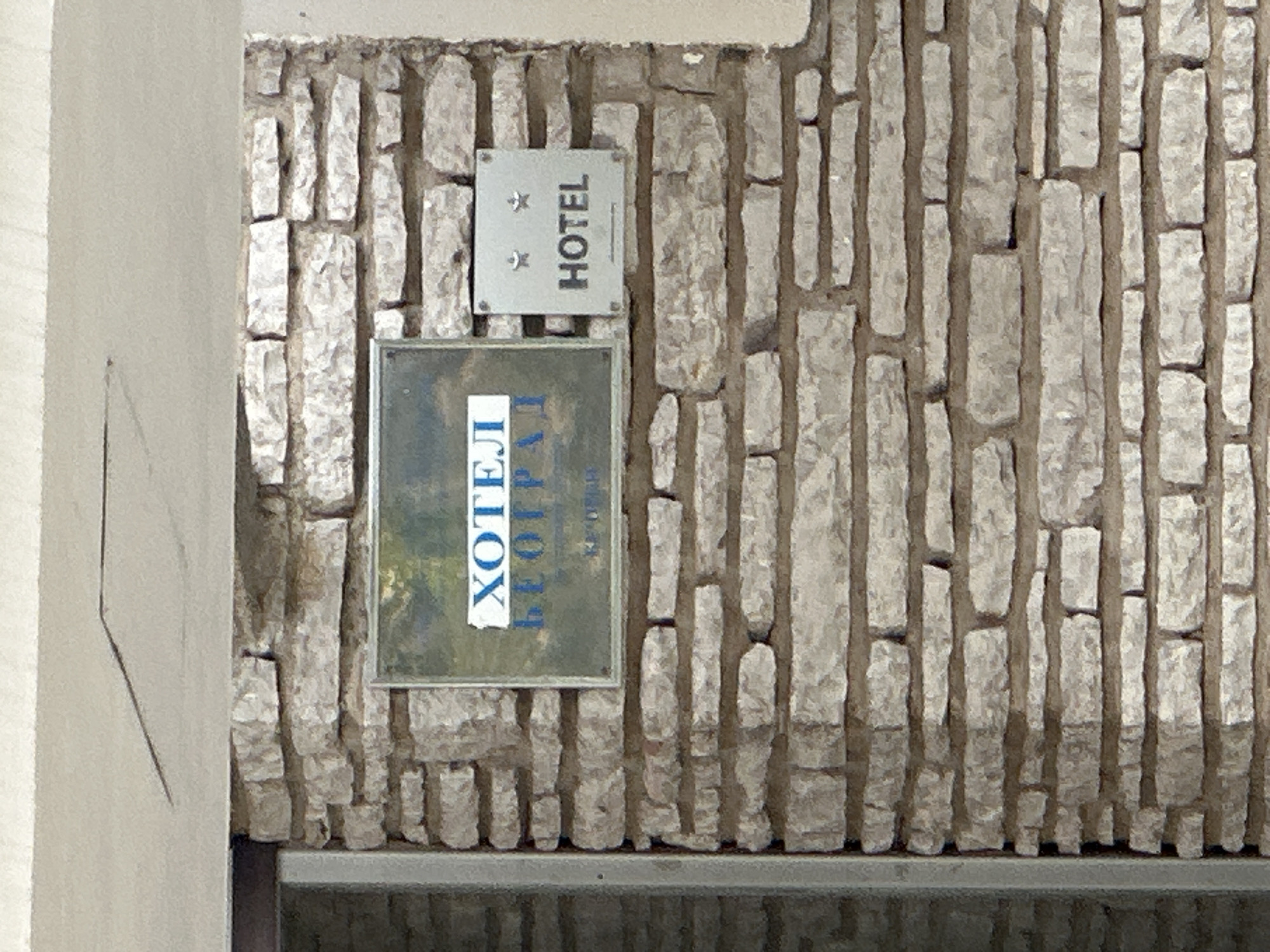 A close-up shows the exterior wall of a hotel in Boreti, Montenegro. The wall is made of rough, light-colored stones, and two signs are affixed to it. The upper sign is square and silver, with the word "HOTEL" printed in black letters and two stylized stars above the word. Below this sign is a larger, rectangular sign with Cyrillic script in blue letters. The signs and the wall are weathered, suggesting a slightly aged or rustic appearance. The bottom part of the image shows a gray surface, indicating either the base of the wall or a ledge in front of it. To the left is a portion of a light-colored wall with a subtle line etched on its surface.