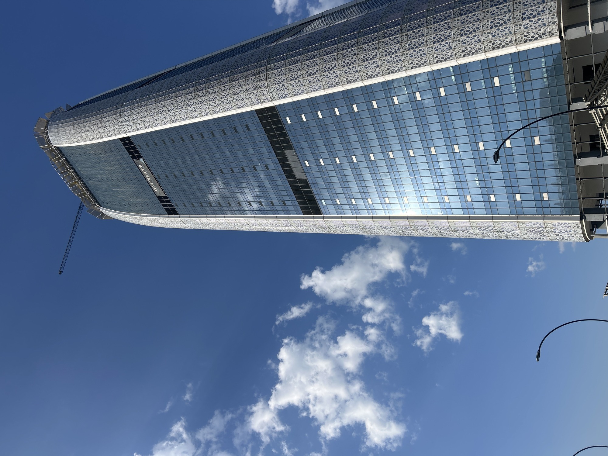 An extreme low-angle, full shot captures a modern, glass-and-metal skyscraper under a bright blue sky with scattered white clouds. The building's facade is a grid of reflective blue glass panels interspersed with smaller white rectangular windows. The right side of the building is adorned with an intricate, white geometric pattern. A construction crane is visible at the very top of the skyscraper, suggesting ongoing construction or renovation. In the lower part of the frame, two black, curved streetlights are positioned against the sky, and parts of a lower structure, possibly an entrance or scaffolding, are visible. The architectural style is contemporary and sleek. The image's perspective emphasizes the immense height and verticality of the building. The overall scene is one of urban development and modern architecture. This building is the Capital Fort building in Sofia, Bulgaria.
