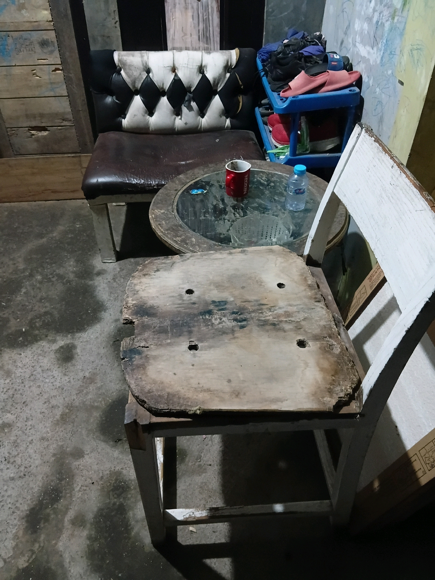 In Cikal, Indonesia, an indoor scene displays a collection of worn and aged furniture. A tufted black and white sofa with a brown leather seat is positioned to the left. In front of it, a circular glass-topped table with a dark wooden frame holds a red mug and a clear plastic water bottle. To the right of the table, a white wooden chair with a distressed wooden seat is visible. Behind the sofa, a blue plastic multi-tiered shelf unit is stacked with various items, including shoes and folded clothing. The flooring is a concrete surface, showing signs of dampness and wear. The background features wooden planks and a light-colored wall with some markings.