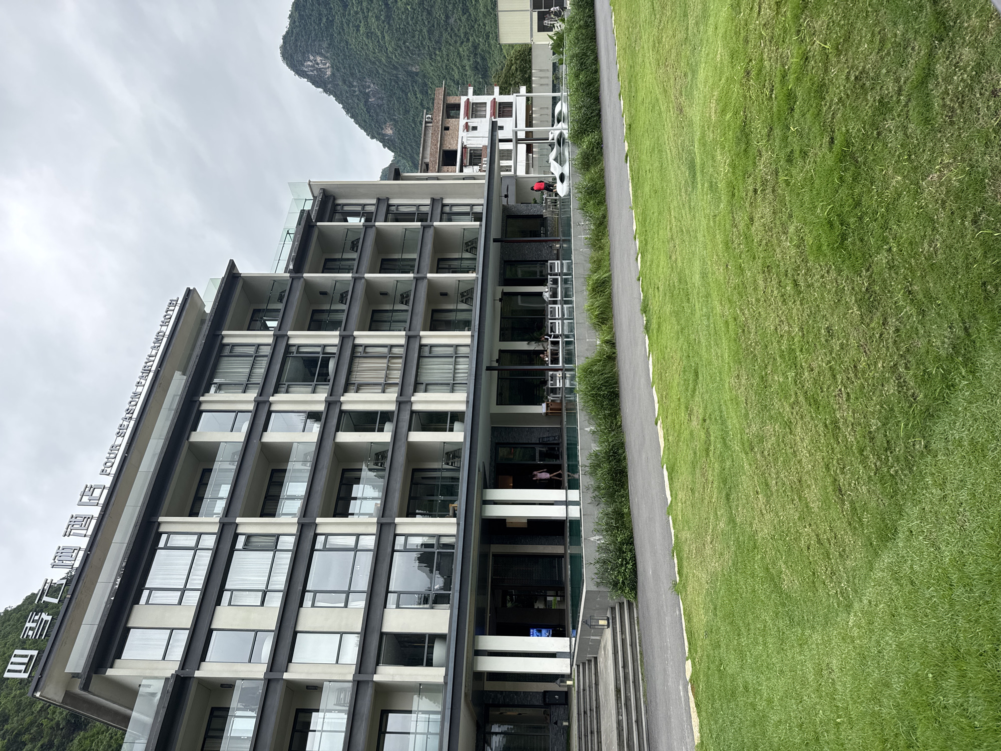 The photograph captures the Four Seasons Fairyland Hotel in Guilin, China, a contemporary architectural structure standing amidst lush greenery and under an overcast sky. The hotel is a multi-story building with a modern facade of glass windows and dark gray structural supports, and it features a distinctive stepped balcony design. The hotel's name is displayed in both English and Chinese characters on the side of the building. Adjacent to the hotel is a well-maintained lawn bordering a narrow paved pathway. In the background, a karst mountain is visible. A smaller building is located further in the background. At least one person can be seen on one of the hotel's balconies. Several cars are parked on the right side of the photograph.