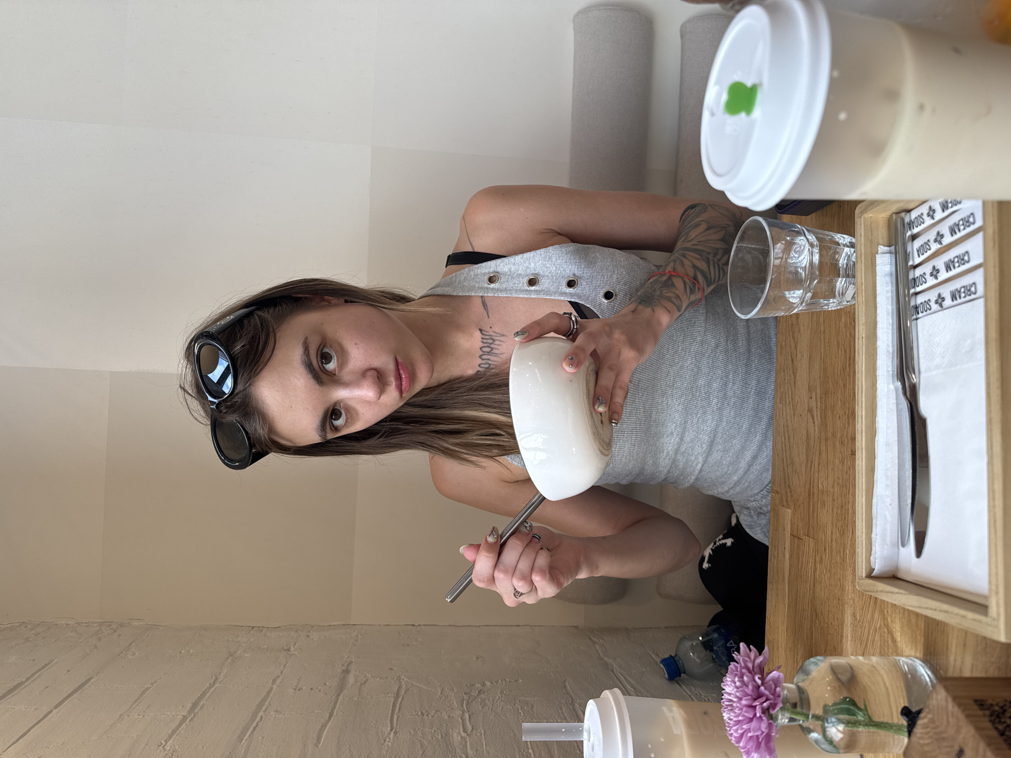 A young woman with dark brown hair, wearing sunglasses perched on her head, a grey tank top, and a black sports bra is seated at a wooden table in a cafe. She is holding a white bowl in her left hand and a metal spoon in her right hand, appearing to be eating. She has tattoos on her chest and arm. On the table, to her left, is a clear plastic cup with a white lid and a straw, and a small glass bottle containing a single purple flower. To her right, there is a clear glass of water and another large clear plastic cup with a white lid. In front of her, on the table, is a wooden tray containing a white napkin and two silver utensils. The background shows a light-colored textured wall and a grey cushioned bench. The overall lighting is soft and natural.