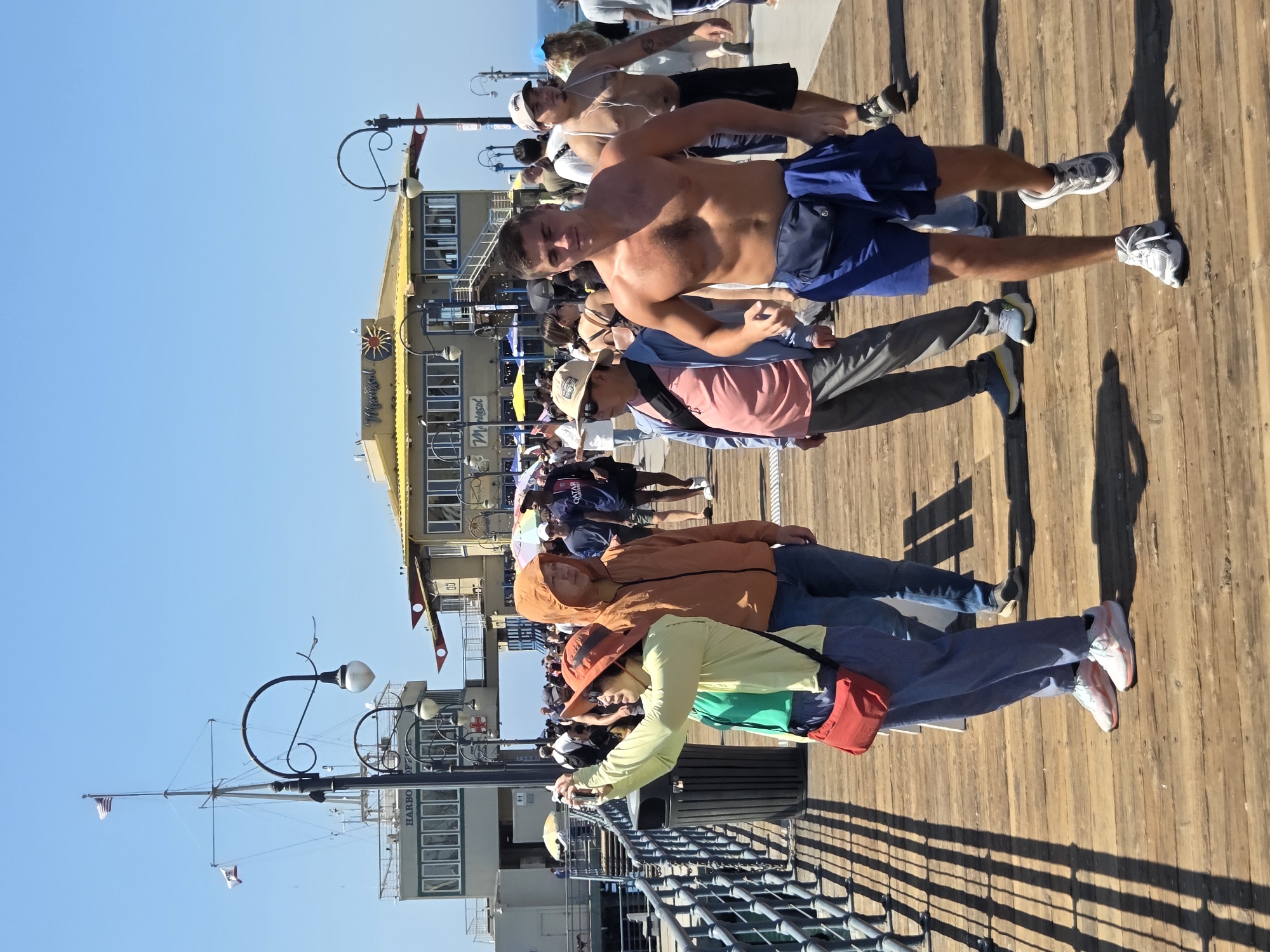 This is a full-shot, eye-level, outdoor, eye-level photograph taken on a sunny day on the Santa Monica Pier in Santa Monica, California. The pier is made of wooden planks and features a railing on the left side. In the foreground, a man with tanned skin and short brown hair is walking towards the camera. He is shirtless, wearing blue shorts with a dark blue fanny pack around his waist and a blue cloth draped over it. He is also wearing white and grey sneakers. To his left, a person wearing an orange hooded jacket and blue jeans is walking away from the camera, with their head turned slightly to the right. Next to them, a woman wearing a light green long-sleeved shirt, blue pants, and white sneakers is holding up a phone to take a picture, wearing a red crossbody bag. She is also wearing an orange hat. Behind them, other people are walking in various directions. In the background, there is a building with a yellow roof and a sign that reads "Marisol." There are also lamp posts, a flagpole with flags, and other structures associated with a pier. The sky is clear and blue.