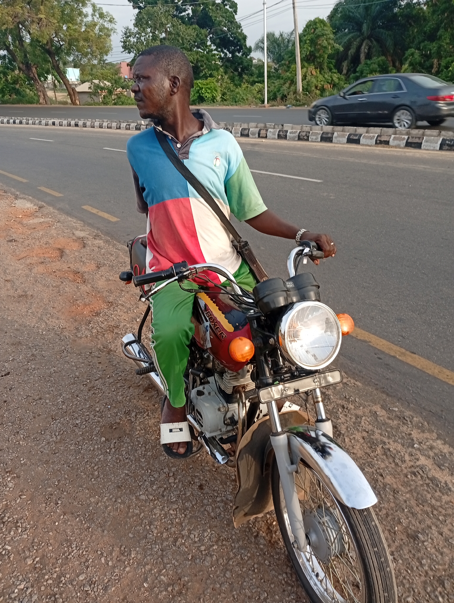 A man sits astride a Boxer motorcycle parked on a dirt shoulder next to a paved road. The man is wearing a short-sleeved shirt composed of blue, white, and red panels, green pants with white stripes, and white sandals. He has a black strap across his chest and is looking to the left. The motorcycle is red and black with a silver front fender and a large headlight. The road has yellow lines and a black and white striped barrier. A car is driving on the road in the background. Trees and buildings are visible further in the background. The scene appears to be in Adankolo, Nigeria.