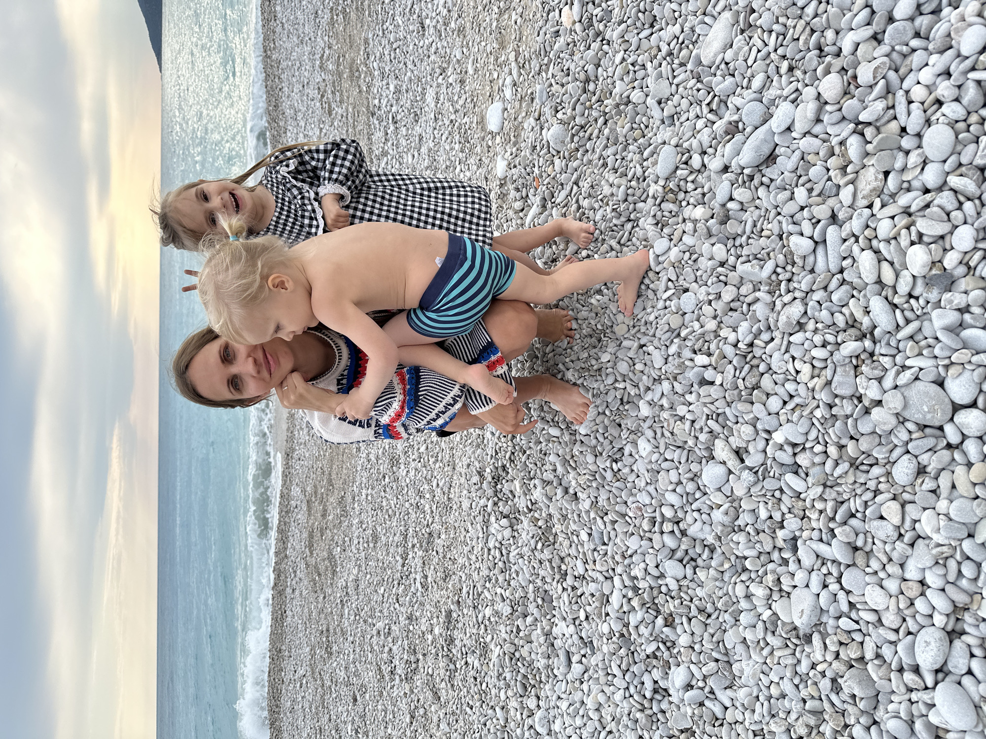 The image captures a candid moment on a pebble beach in Fethiye, Turkey. A woman kneels, supporting a young child in blue and green striped swimwear on her lap. An older child wearing a black and white checkered dress is positioned behind the woman. The backdrop features the turquoise water of the Mediterranean Sea meeting the pebble beach. The sky is overcast with light clouds.