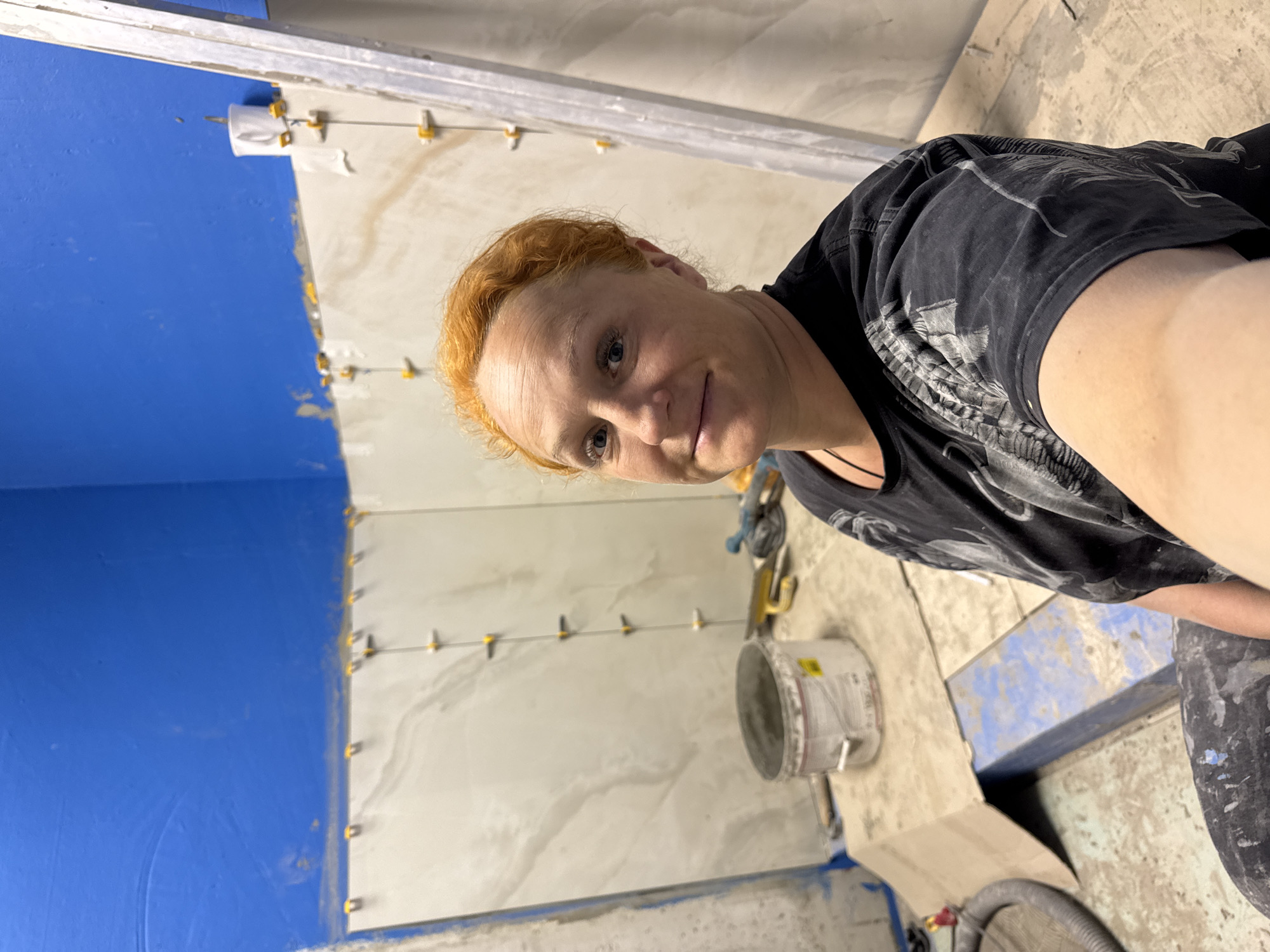 A person with red hair is taking a selfie in a room under construction. A blue wall is visible on the left. White marble-like tiles are being installed on the walls. There are yellow leveling wedges between the tiles. A bucket and tools are on the floor. The specific location in Chaikovsky, Russia, cannot be determined from the image.