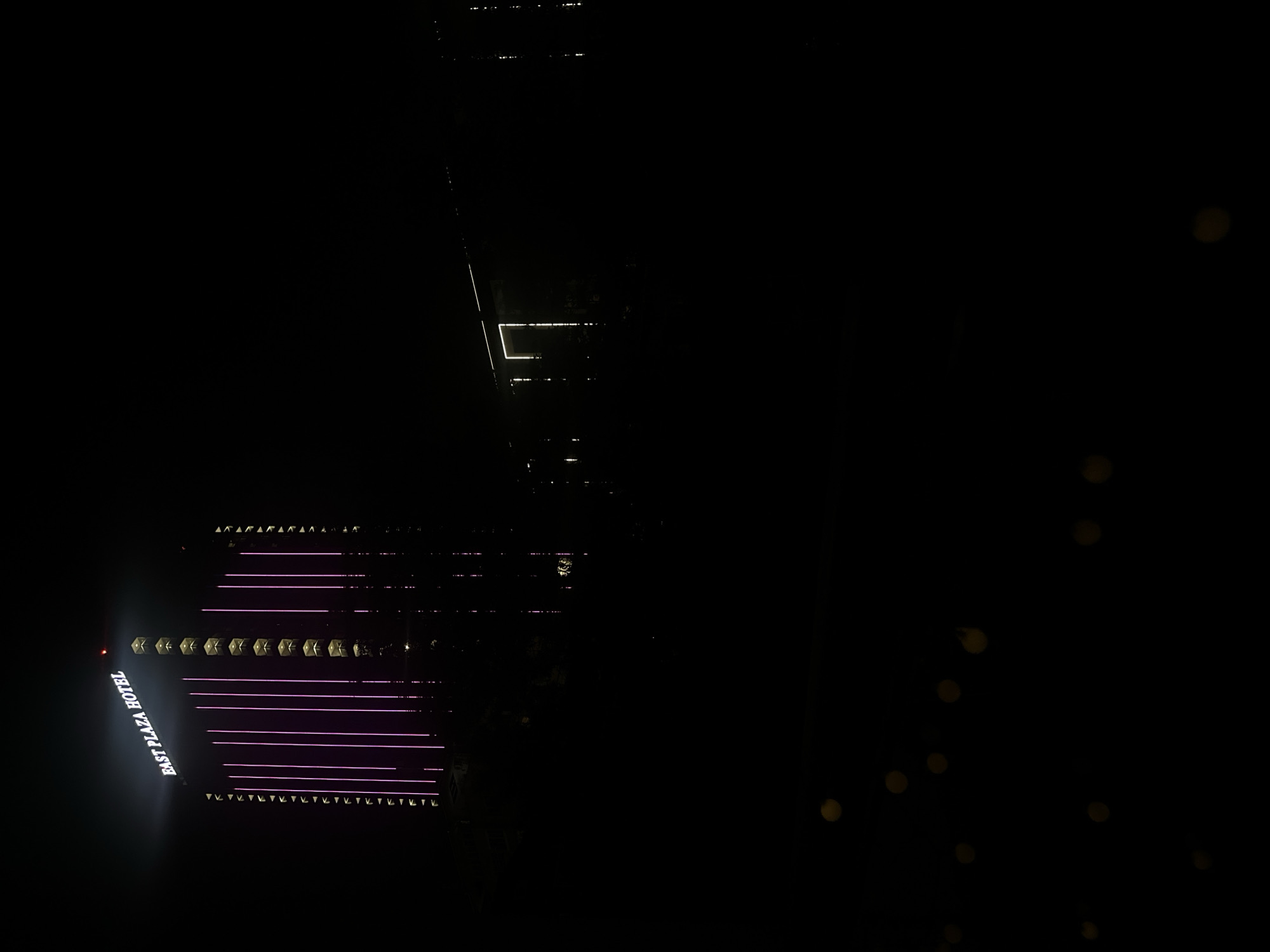 The image is a nighttime exterior shot of the East Plaza Hotel in Sofia, Bulgaria. The tall, modern hotel is illuminated with vertical pink lights along its facade, and the hotel name is visible in white lettering at the top. To the right of the main hotel building, a lower, darker structure with a large rectangular outline of lights is visible. The foreground is almost entirely black, with some out-of-focus circular lights that appear to be string lights or bokeh effects from distant lights. The overall scene depicts the hotel illuminated at night, with surrounding darkness.