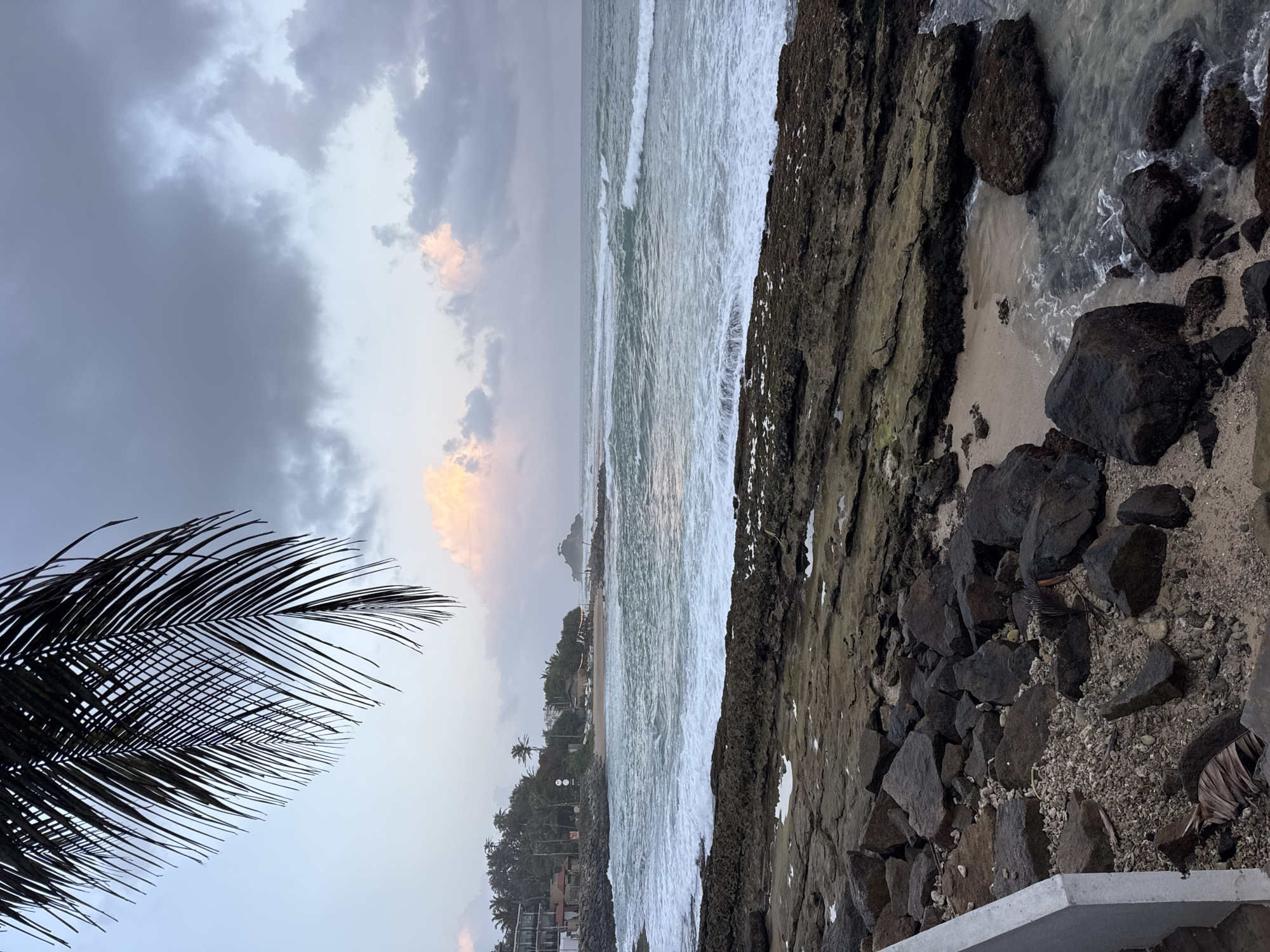 This is a photograph of the coastline in Goviyapana, Sri Lanka, at either dawn or dusk. In the foreground, a rocky shore is visible, with large, dark, irregularly shaped rocks interspersed with smaller pebbles and sand. The water is a greenish-blue, with white foam from waves crashing against the rocks. In the midground, a sandy beach is lined with palm trees and several buildings, including what appear to be hotels or guesthouses. Beyond the beach, the ocean stretches to the horizon, with a small, rocky island visible in the distance. The sky is overcast with shades of grey and purple, with patches of soft pink and yellow light indicating the rising or setting sun. A large frond of a palm tree hangs from the top left corner of the frame, partially obscuring the sky. There are no people visible in the image.