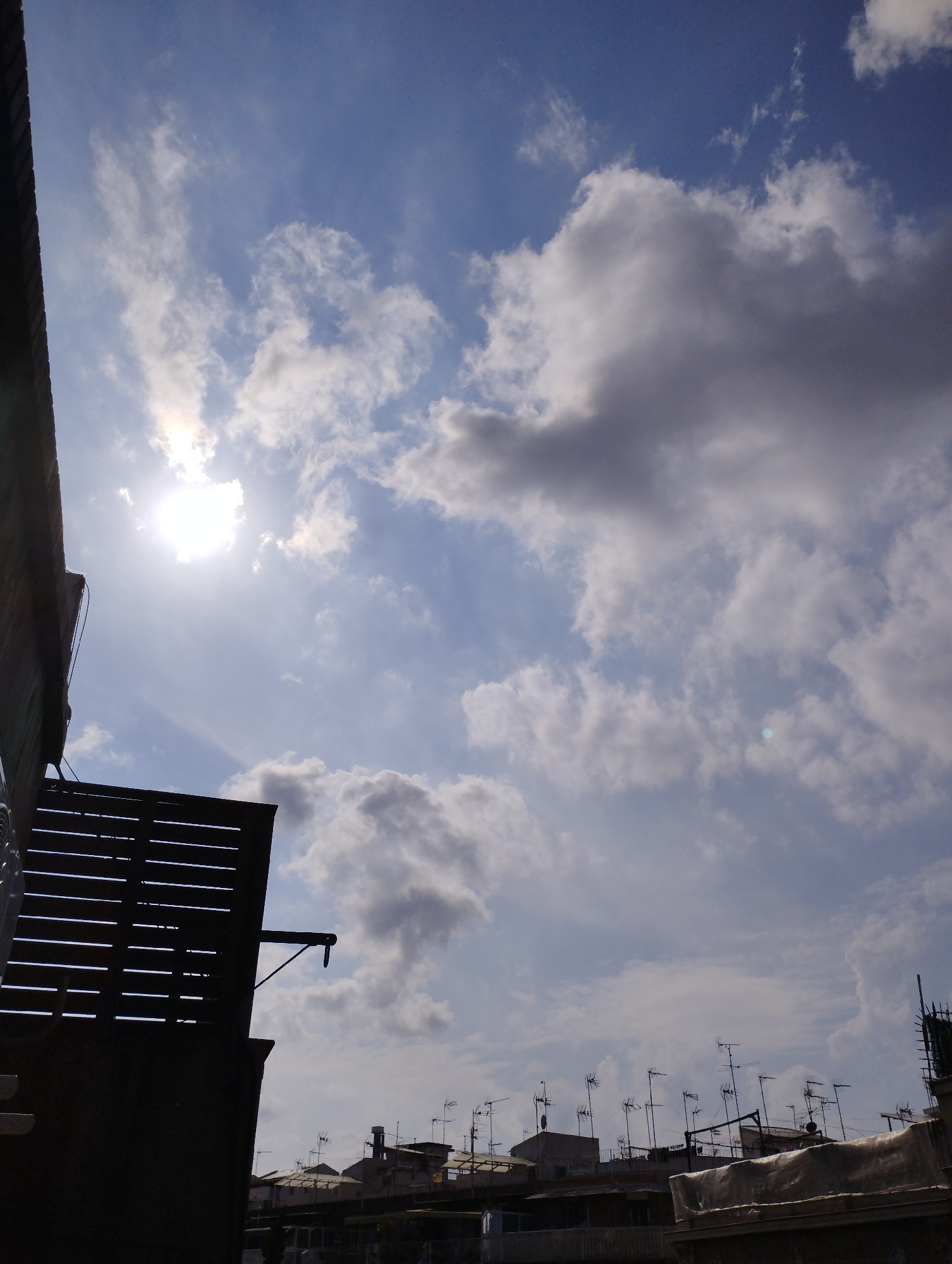 The image presents an upward-looking view of a partly cloudy sky over an urban or suburban landscape during the daytime. A bright, intense sun dominates the upper-left quadrant, appearing as a strong glare and partially obscured by thin clouds, backlighting the entire scene. The sky varies from a lighter blue near the sun to a deeper azure, filled with numerous clouds ranging from wispy white formations to larger, denser cumulus or stratocumulus clouds that appear grey and voluminous.
In the foreground on the far left, a dark, heavily silhouetted building structure, possibly a wall or part of a roofline, extends vertically. Below this, a distinctive dark, slatted structure, likely a balcony railing or a decorative screen, extends horizontally into the lower-left quadrant.
Along the horizon line in the lower portion of the image, the rooftops of multiple buildings are visible. These rooftops are mostly silhouetted against the brighter sky and are characterized by a dense array of television or radio antennas, protruding upwards from various points. Other indistinct rooftop features, including what appears to be covered objects or small structures, can also be seen.
No people are visible, and no specific event or activity is taking place; the image captures a static moment of the environment. The time of day is clearly daytime, indicated by the high and bright sun. The weather is sunny with varying cloud cover. The numerous antennas on the rooftops strongly suggest a residential urban or densely populated suburban setting. There is no visible text in the image.