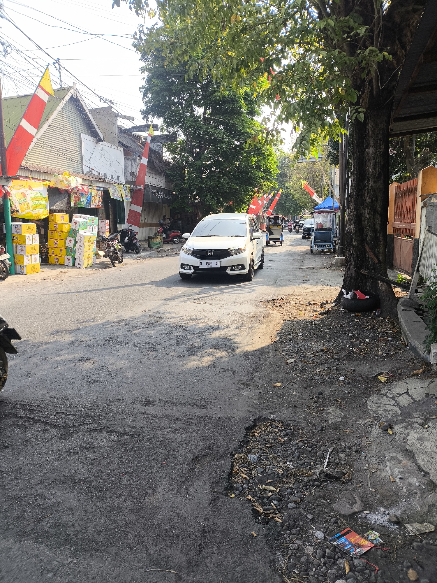 This image depicts a street scene in Lumajang, Indonesia, likely during daytime. The street is paved with asphalt and shows signs of wear and tear, including several potholes and cracks. On the left side of the street, there are several storefronts with displays of goods, including boxes of what appear to be beverages or food items. Red and white flags, possibly indicating a celebration or national holiday, are visible on poles and attached to buildings. A white Honda Mobilio with the license plate N 1894 ZT is driving on the road. Several motorcycles and what appear to be motorized tricycles (becak motor) are also present, some parked and others in motion. A large tree with dense foliage stands on the right side of the street, casting shadows. The environment appears somewhat cluttered, with debris and trash visible on the side of the road, including a discarded tire and scattered litter. The overall impression is of a busy, lived-in urban street.