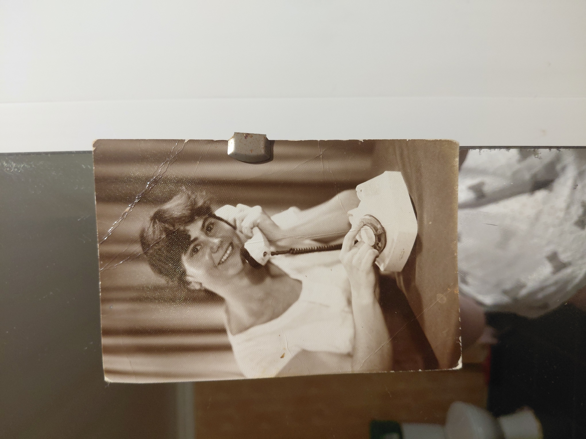 The image is a sepia-toned photograph of a young woman holding a rotary telephone to her ear. She is smiling and looking towards the viewer. The woman is wearing a light-colored, possibly white, dress with a rounded neckline. The telephone is white and appears to be a desk model. The background behind the woman consists of vertical blinds or drapes. The photograph is positioned on a surface, and a portion of another image or object is visible at the bottom, appearing to be a white garment with a pattern. A metallic object is attached to the right edge of the photograph, possibly a clip or magnet. The photograph itself shows signs of age, with creases and light damage. The image is framed by a white background and a darker area to the left. No specific location in the city of Marynivka, Ukraine can be identified from this photograph.