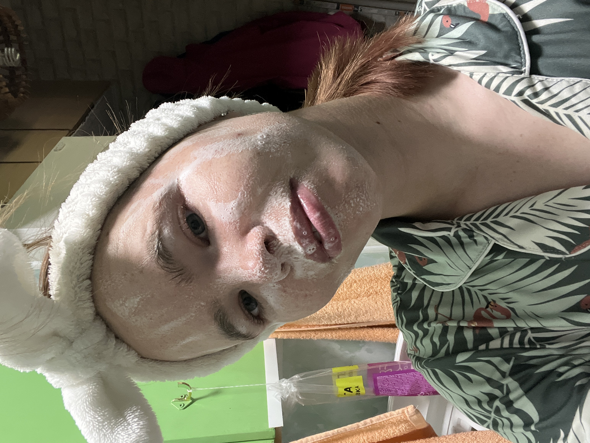 A close-up, selfie-style shot shows a young person with a white, fluffy headband with ears on it, appearing to be in a bathroom. The person's face is covered in a white, foamy substance, likely a face mask or cleanser. They are wearing a green pajama top with a tropical leaf and flamingo pattern. In the background, there are green cabinets, orange towels hanging on hooks, and a plastic bag with yellow and purple packaging. The image is taken from a slightly upward angle, focusing on the person's face. The overall scene suggests a personal grooming or self-care moment. The provided context states the location is Chaikovsky, Russia.