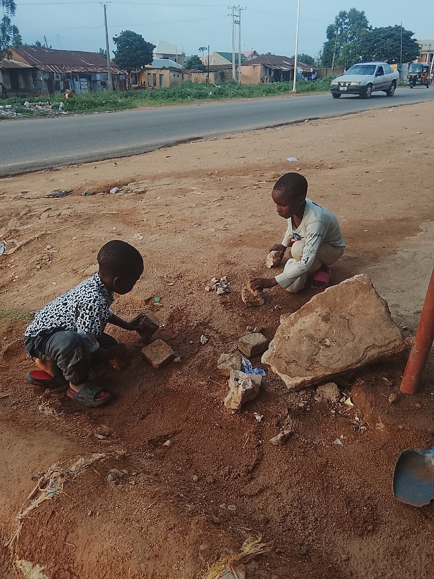 Two young children are playing on a dirt patch adjacent to a paved road in Vom, Nigeria. One child, wearing a patterned shirt and dark pants, is kneeling and appears to be arranging or manipulating small rocks or bricks. The other child, wearing a light-colored long-sleeved shirt and pants, is also kneeling and engaged with similar small rocks. A large rock is nearby, as is a shovel. The road is in the background, with buildings, foliage, and a car and a motor tricycle visible. The time of day seems to be daylight.