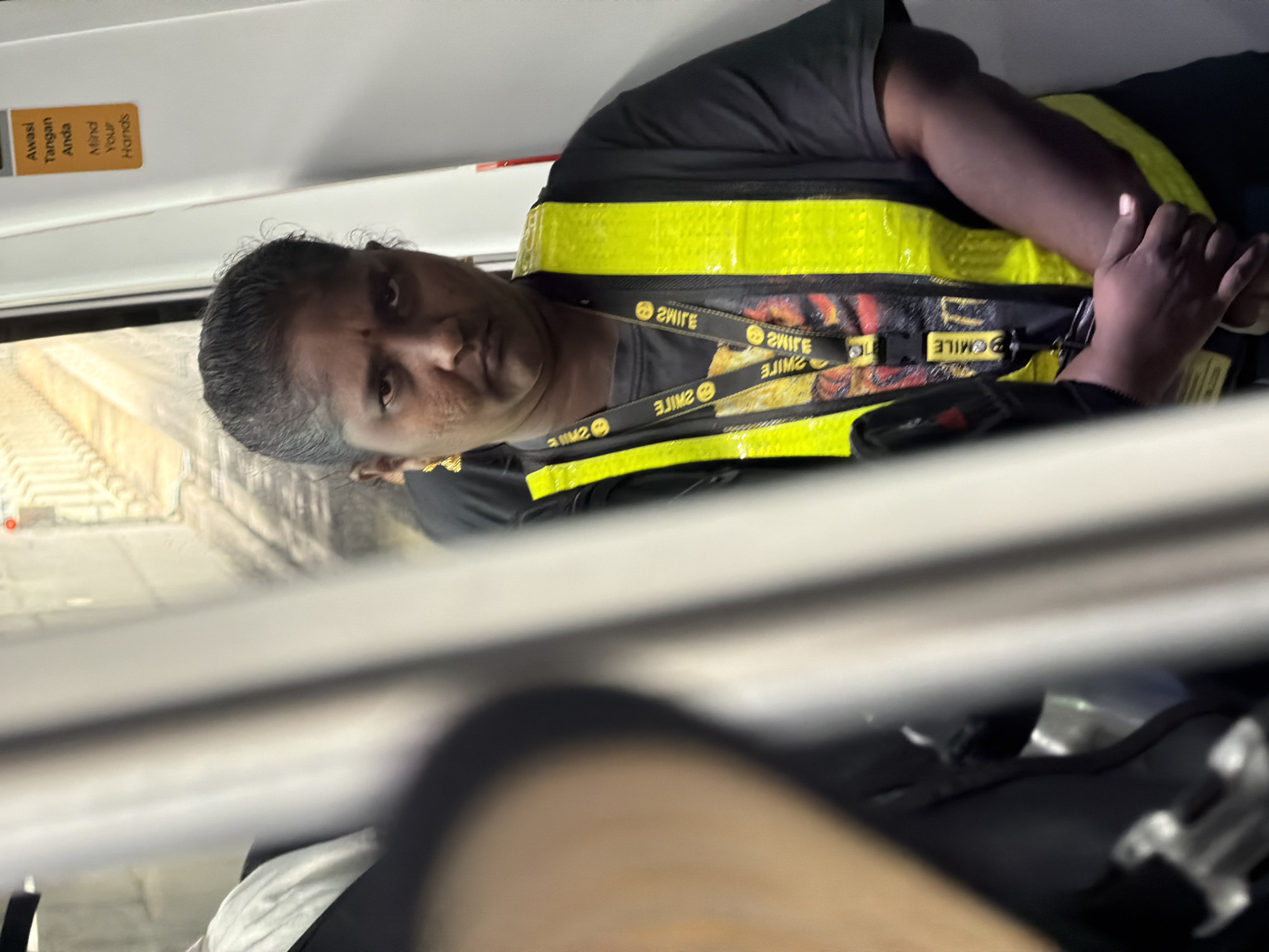 A person with dark skin and dark hair pulled back is standing indoors, likely on a train, wearing a black t-shirt and a bright yellow reflective vest. They have a stern expression and are wearing gold earrings. Around their neck, they have lanyards with the word "SMILE" printed on them. There is a sticker on a white surface to the right of the person that says "Awasi Tangan Anda" and "Mind Your Hands" in English. The background appears to be a train interior with visible tracks and lighting.