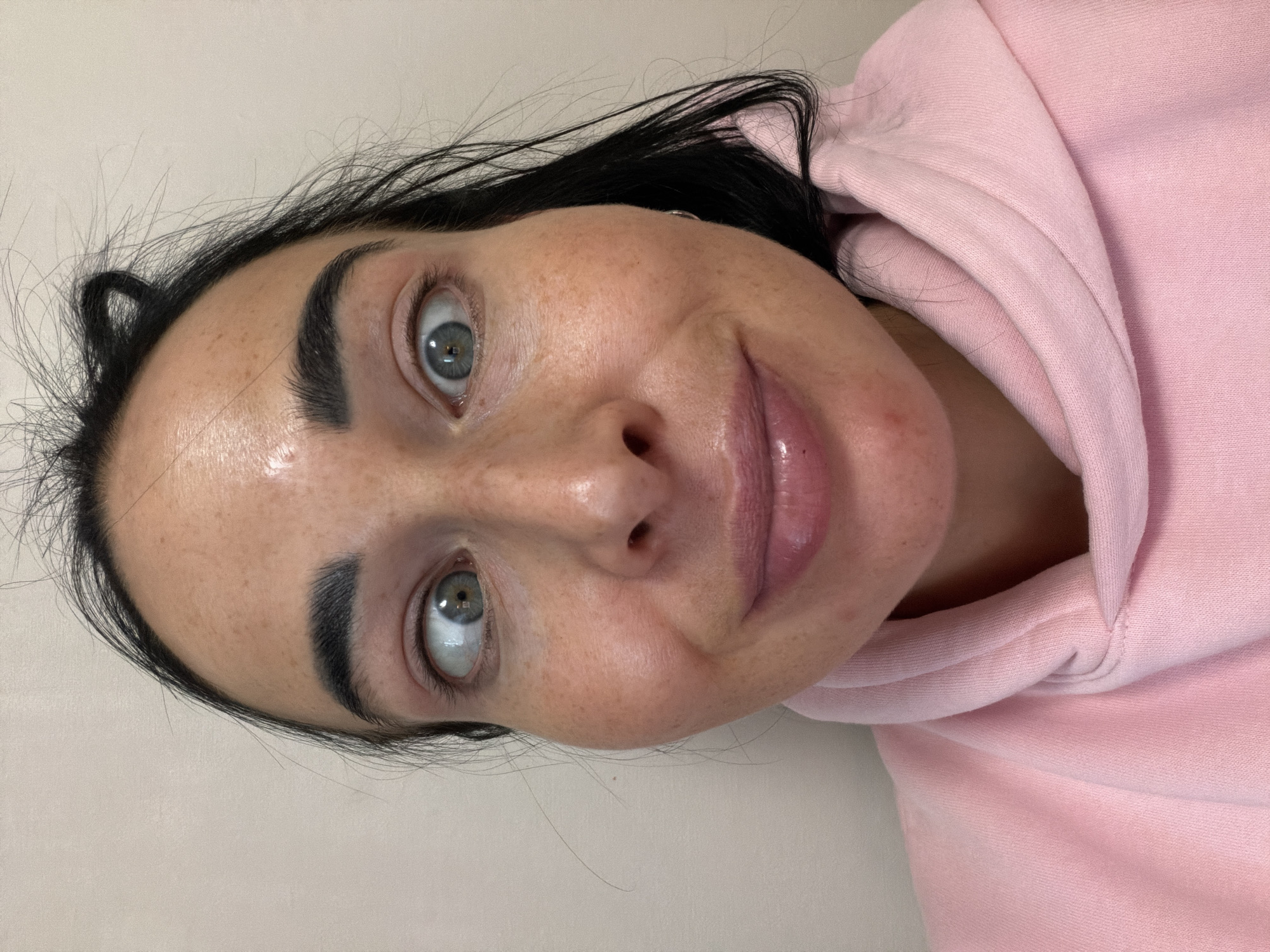 This close-up portrait, rotated 90 degrees counter-clockwise, features a woman looking directly at the camera. She has long, dark, slightly dishevelled hair and fair skin adorned with numerous freckles across her forehead, nose, and cheeks. Her prominent, dark eyebrows frame striking light blue or green eyes, which are wide open, conveying an intense and direct gaze. Her full lips are slightly parted and appear glossy, with a subtle, ambiguous expression that could be interpreted as a slight smirk or hint of a smile.
The woman is wearing a comfortable-looking, light pink hooded sweatshirt (hoodie), parts of which are visible on the right side of the frame. The background is a plain, light-colored surface, suggesting an indoor setting with even lighting. There is no visible text within the image. While the location context of Saint Petersburg, Russia, is provided, there are no specific visual cues in the image to confirm this.