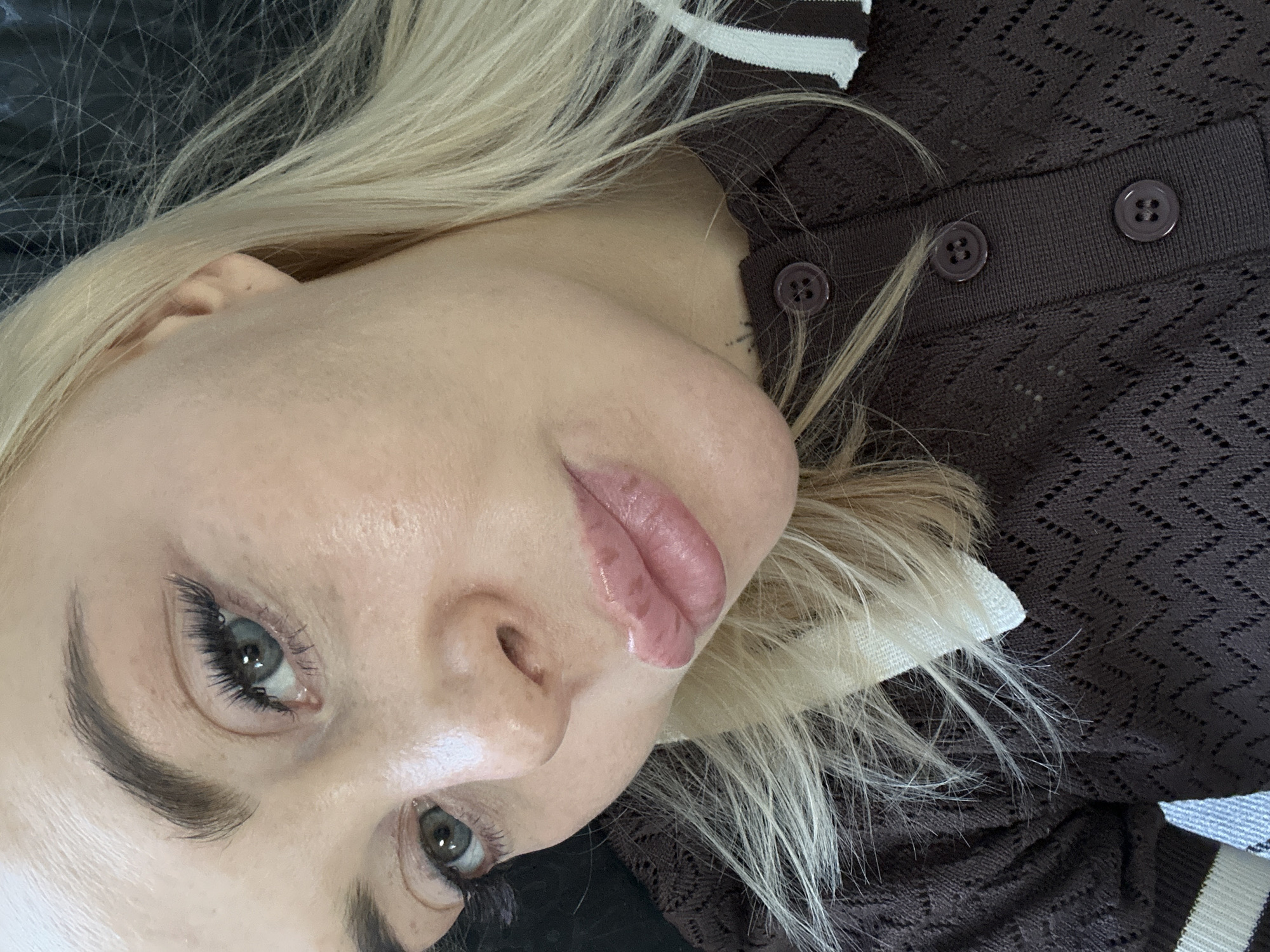 A close-up, high-angle portrait features a young woman with fair skin and long, straight blonde hair, lying down and looking towards the viewer. Her head is tilted, with her face occupying the left side of the frame. She has prominent, neatly shaped dark eyebrows and long, dark eyelashes framing her green or blue eyes. Her lips are full and appear to have a glossy, light pink or nude colored product on them. Her skin shows a natural, slightly dewy texture.
She is wearing a dark brown knitted top or cardigan with a patterned texture and visible round, brown buttons along a placket. There are also white and light blue or grey stripes visible on a collar or trim of the garment on the right side of the image. Her blonde hair is spread out around her head, contrasting with the dark brown of her clothing and a dark, possibly textured, background surface on the top left. The overall impression is a calm, natural portrait with a focus on facial features. There is no visible text in the image.