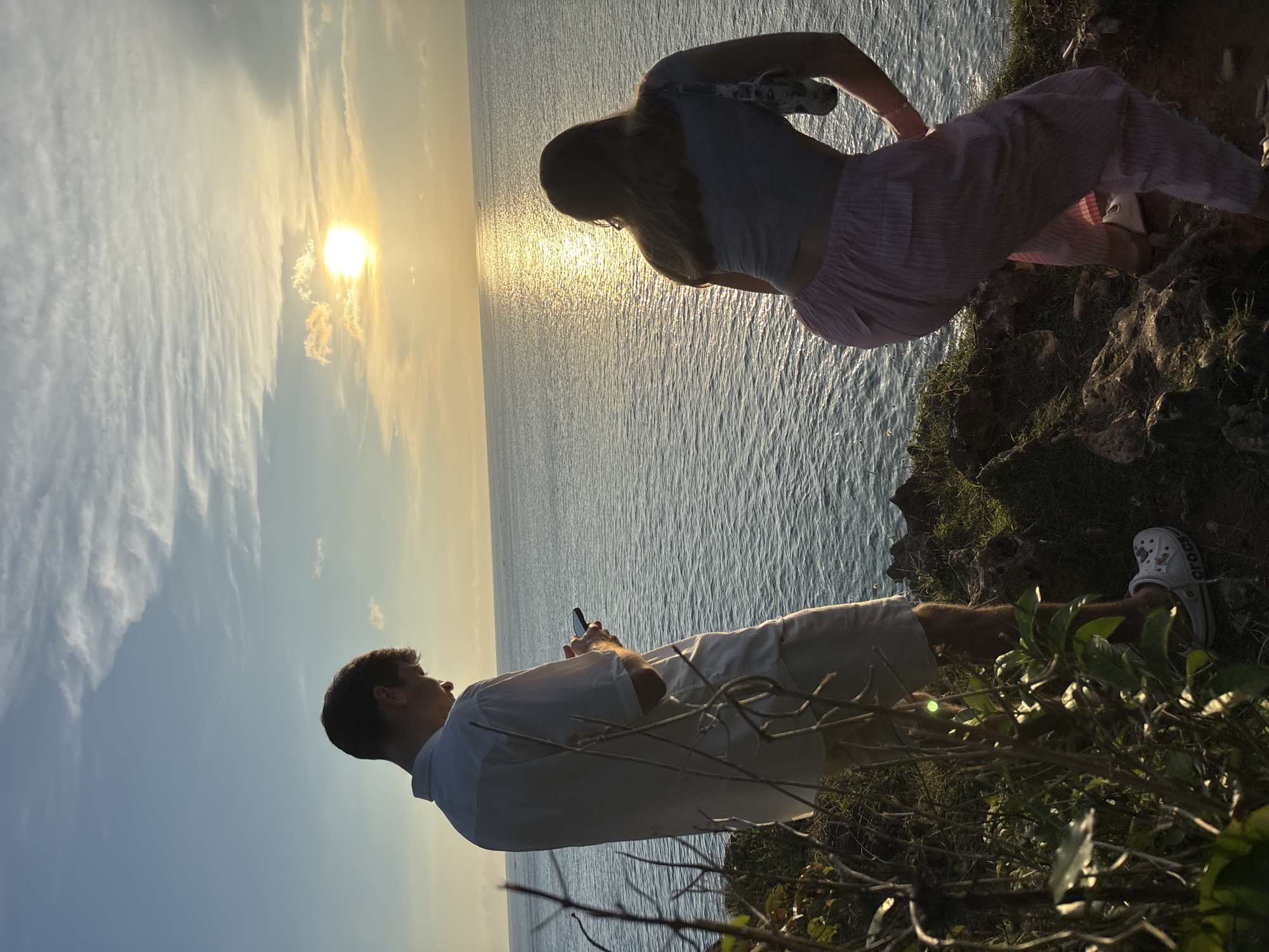 A man and a woman are standing on a rocky cliff edge overlooking the ocean in Kuta Selatan, Indonesia. The man is standing, wearing a white shirt and light-colored shorts, looking at his phone. The woman is leaning over the cliff edge, wearing a light-colored top and pink striped pants. The sun is setting over the ocean, creating a bright reflection on the water. There are bushes and foliage around the cliff edge. The man is wearing white Crocs.