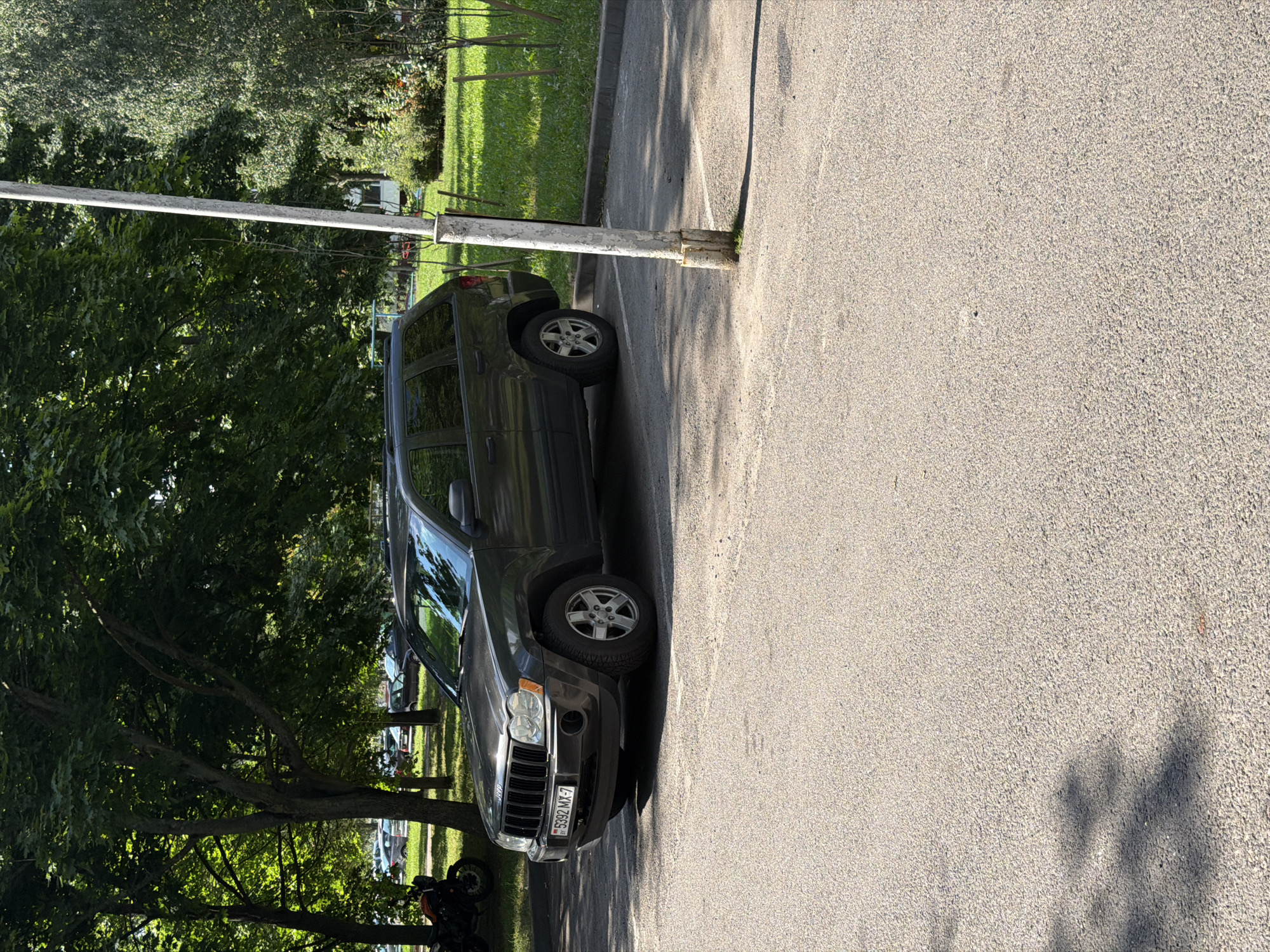 The image depicts a parking lot in Minsk, Belarus, on a sunny day with shadows from trees cast across the asphalt. A dark gray Jeep Grand Cherokee is parked in the foreground, facing left. The license plate of the Jeep reads "5392 MX-7". To the left of the Jeep, partially visible, is a motorcycle. The background features a grassy area with trees and a white pole. The overall scene is of a typical urban parking area.