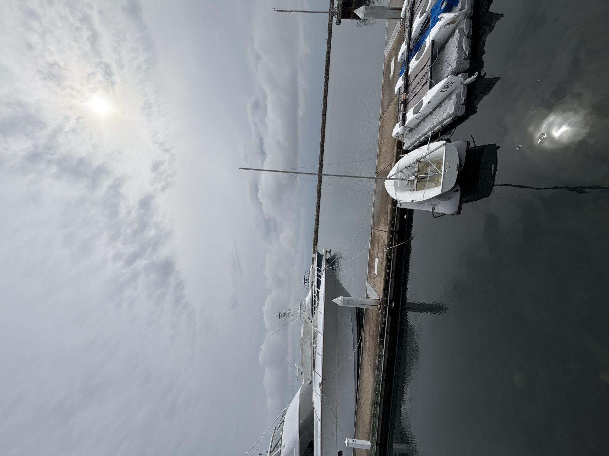 The image depicts a marina with several boats docked. In the foreground, a small white sailboat with a tall mast is moored. To its right, a line of kayaks or small boats, some covered with tarps, are also docked. In the background, larger white yachts are visible, tied to a dock. The sky is overcast with patches of sunlight breaking through the clouds, creating reflections on the water. A stone breakwater extends into the distance. The scene appears to be set in a coastal area, likely a marina in Veracruz City, Mexico, given the context. There are no people visible in the image.