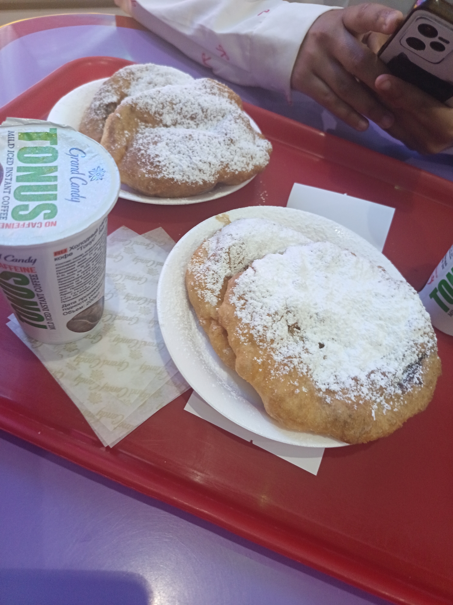 The image shows a close-up of a red tray on a table with a purple surface. On the tray, there are two white plates, each containing a pastry dusted with powdered sugar. To the left of the plates is a white cup with a label that reads "TONUS MILD ICED INSTANT COFFEE NO CAFFEINE". The cup also has "Grand Candy" printed on it, along with some text in Cyrillic. A person's hands are visible in the upper right corner of the image, holding a smartphone. The overall scene appears to be a casual dining setting, possibly a cafe or restaurant, where someone is enjoying a snack and a drink while using their phone. The lighting suggests it could be daytime, but there are no strong indicators of time of day or weather. The focus is on the food and drink items, with the person's activity being secondary.