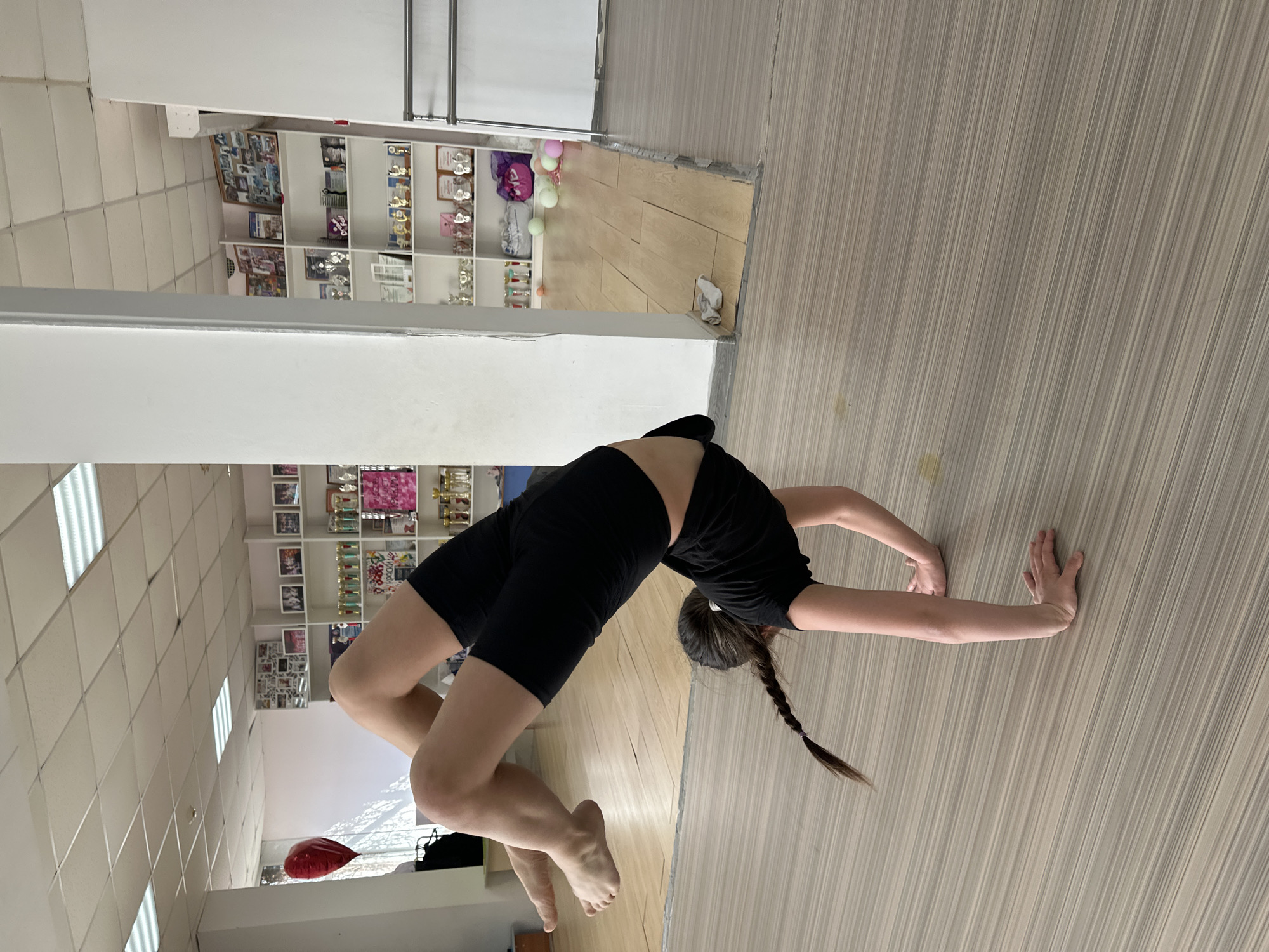A young person performs a backbend (also known as a bridge pose) on a light-colored wooden floor in an indoor setting. The individual, likely female given the long, dark braided hair, is dressed in a black short-sleeved top and black athletic shorts, and is barefoot. They support their arched body with their hands and feet flat on the floor, showcasing flexibility.
The indoor environment appears to be a studio or practice room. The floor beneath the person is made of light-toned wooden planks. To the right of the person, the floor transitions to a light grey, vertically striped laminate, with a small crumpled white item (possibly a sock) situated where the two floor types meet.
In the background, a white wall is adorned with multiple open shelving units. These shelves are filled with numerous small golden and silver trophies or awards, alongside framed pictures and certificates, suggesting a space for accomplishments. Some colorful balloons (pink, green, and white) are visible on shelves in the upper background. A large mirror on the far wall reflects parts of the room. The ceiling is white and tiled, featuring rectangular fluorescent light fixtures that provide bright, even lighting. A red balloon is partially visible in the lower left corner of the frame. There is no discernible text in the image.