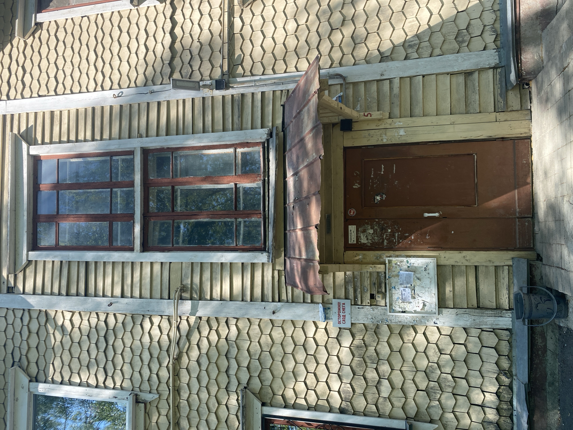 The image shows a two-story building in Novouralsk, Russia. The building has a distinctive facade, composed of hexagonal wooden shingles on the first story and vertical wooden planks on the second story. A window with a brown frame is visible on the left. A brown door is to the right with a piece of rusty metal serving as an awning above the door. Below the door is an aging bulletin board with tattered notices. A sign reading "ОСТОРОЖНО! СХОД СНЕГА!" (Caution! Snow Falling!) is posted next to the bulletin board. A bucket sits next to the building at the base.