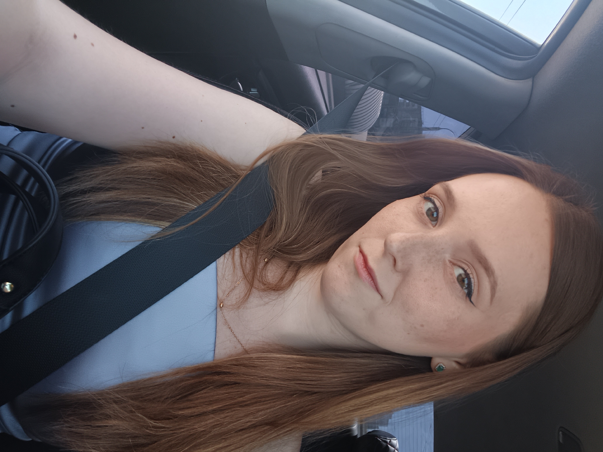 A woman with long, auburn hair is sitting in the driver's seat of a car, looking at the camera. She is wearing a light blue strapless top and a black seatbelt. She has freckles on her face and is wearing a small, green earring. The interior of the car is visible, including the dashboard and a portion of the passenger seat. The background shows a blurry outdoor scene, suggesting the car is either parked or moving slowly.