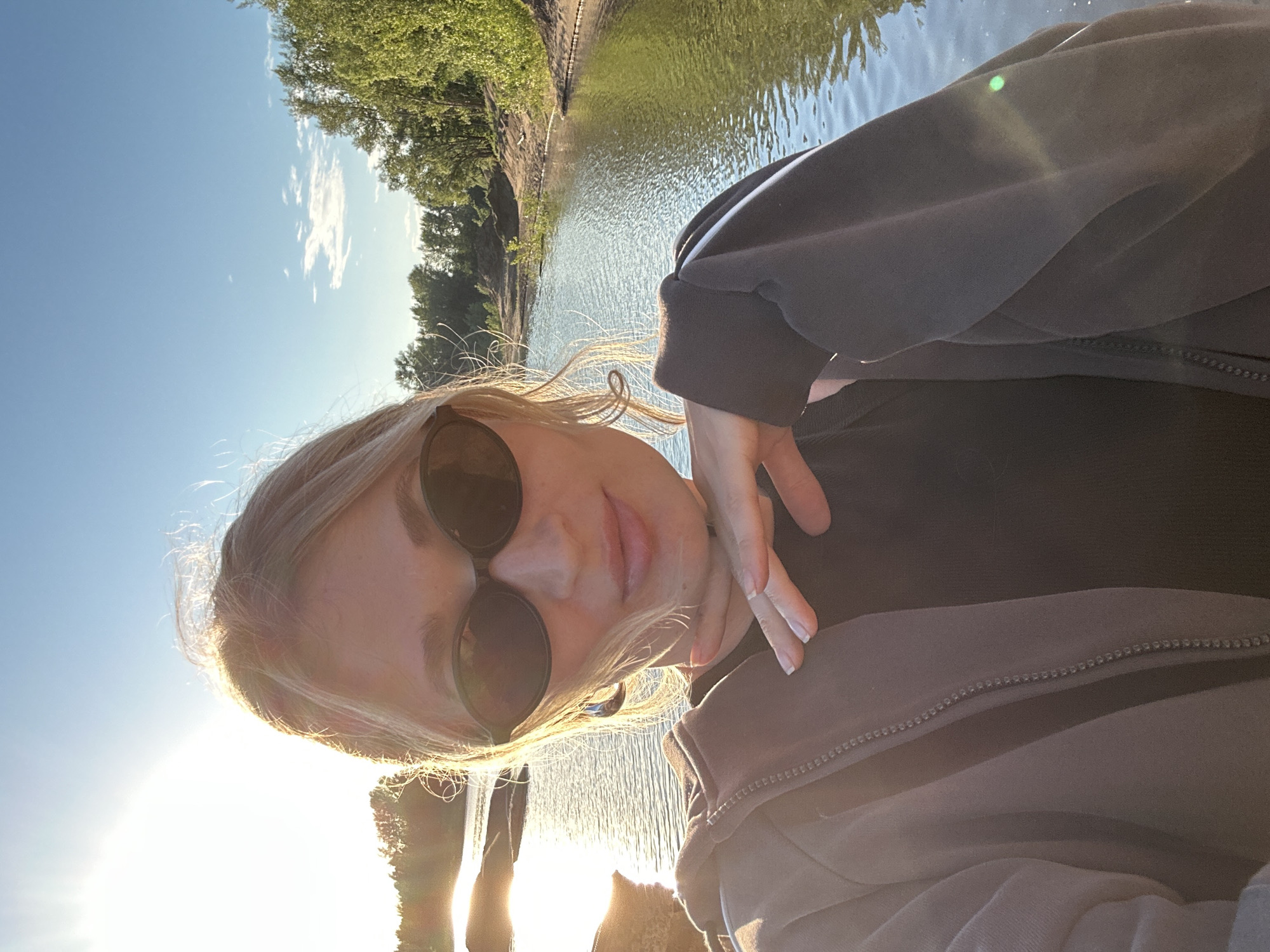 A young woman with blonde hair is taking a selfie outdoors by a body of water. She is wearing sunglasses, a dark top, and a grey zip-up hoodie. Her hand is resting on her chin. The sun is setting, casting a warm glow on the scene and creating lens flare. Trees and a sandy bank are visible in the background. The location is likely the Kama River near Chaikovsky, Russia, a popular spot for recreation.