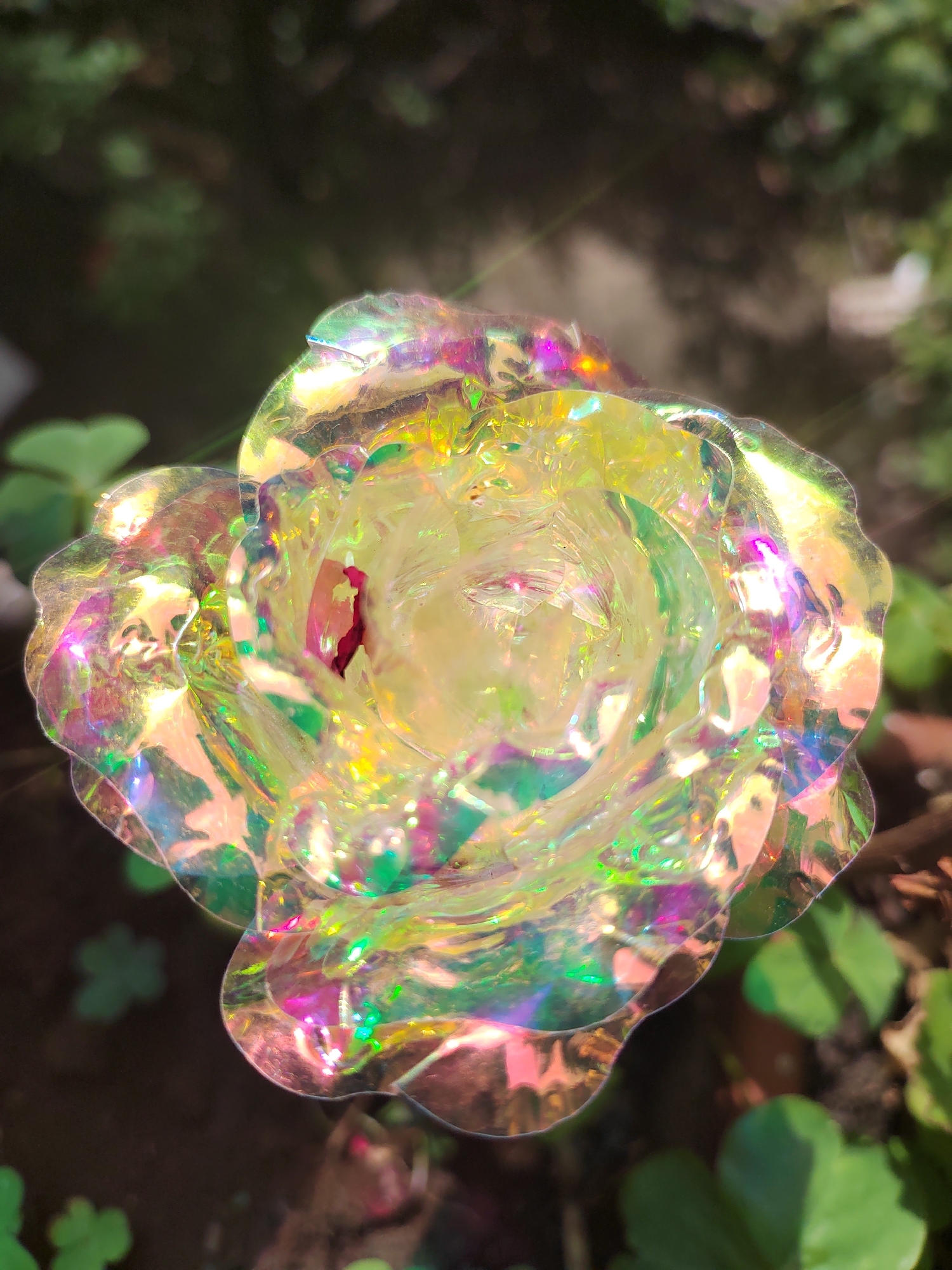 This is a close-up, top-down shot of a decorative, iridescent rose. The rose appears to be made of a translucent, iridescent material that refracts light, creating a spectrum of colors including yellow, green, pink, and purple. The petals are layered and textured, with a central core that has a slightly darker, reddish hue. The rose is positioned outdoors, with a blurred background of green foliage and dark earth, suggesting a garden or natural setting. The lighting indicates it is daytime, with sunlight highlighting the rose's shimmering surface and creating lens flare effects. The shallow depth of field isolates the rose, making it the sole prominent subject. There is no visible text in the image.