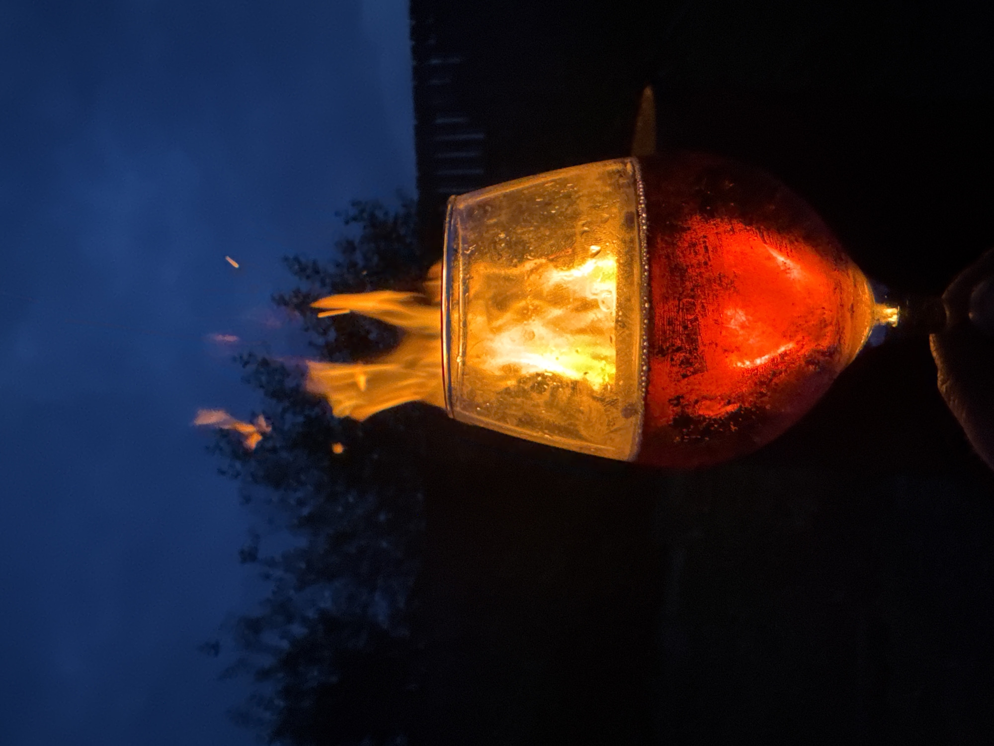 A close-up shot captures a glass of dark red liquid, possibly wine or brandy, in the foreground. Flames are visible inside the glass, creating a dramatic visual effect. The glass is set against a dark, out-of-focus background that suggests an outdoor setting at dusk or night, with hints of trees and a fence. The sky is a deep blue. The lighting is primarily from the flames, casting a warm glow on the glass and its contents. No specific location in Chaikovsky, Russia, is identifiable from the image.