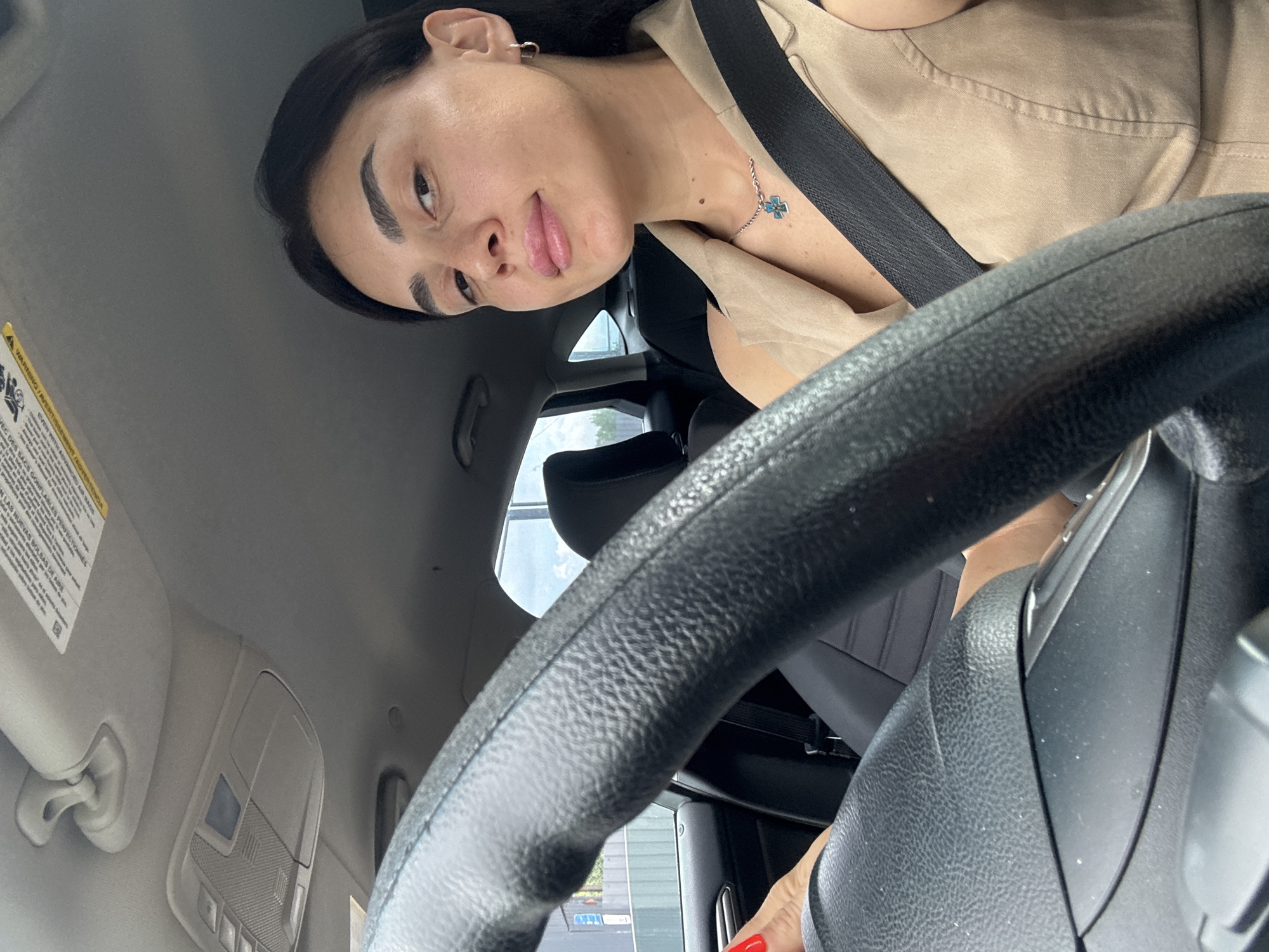 The image shows a woman driving a car. She is wearing a tan sleeveless top and a necklace with a cross-shaped pendant. She has a seatbelt on. The steering wheel is in the foreground. The interior of the car is visible, including the sun visor with labels and the overhead console. Outside the car, a building and a fence can be seen. The sky is cloudy. The specific location in Tbilisi, Georgia, cannot be determined from the image.