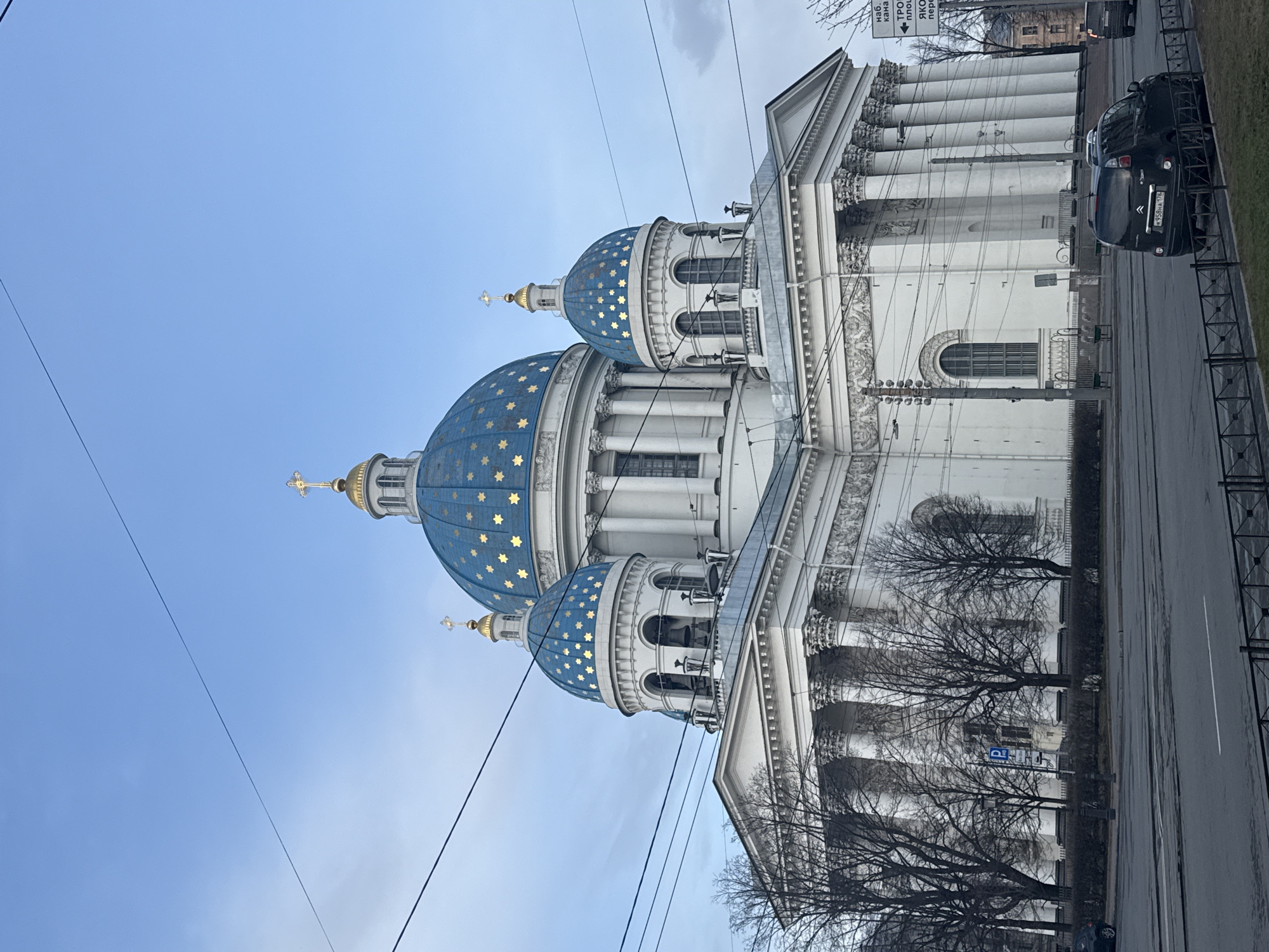 The image shows the Trinity Cathedral in Saint Petersburg, Russia, a large white and blue baroque-style cathedral with five domes. The domes are blue with gold stars, and the cathedral has many white columns. The photo was taken at street level, with cars parked along the road. Power lines are visible in the sky. Bare trees stand in front of the building. A street sign is present.