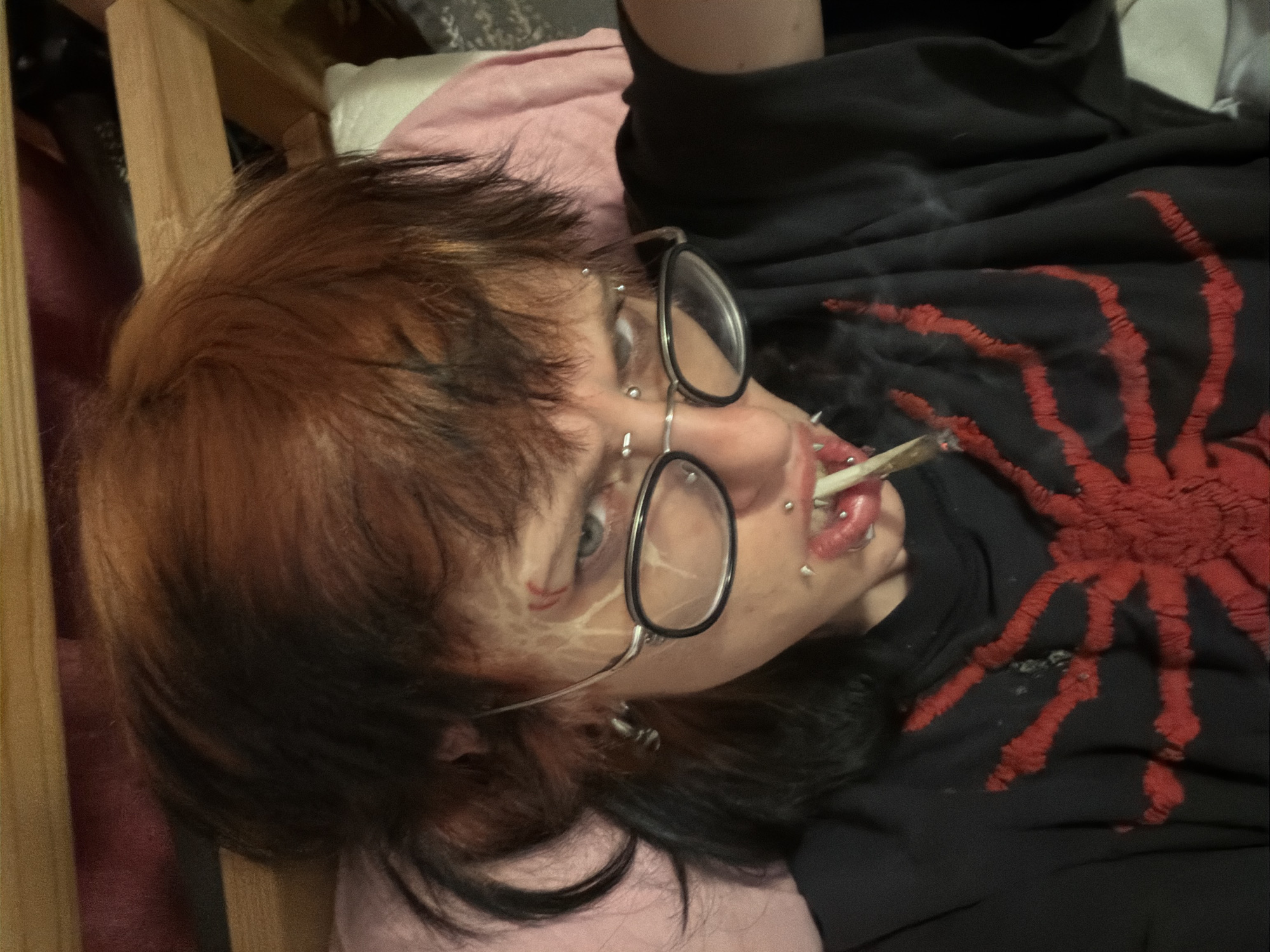 A person with dark hair and reddish-brown highlights, wearing round glasses and multiple facial piercings, is lying down and holding a smoking object in their mouth. The person is wearing a black shirt with a large red spider design. The person is on a pink surface and appears to be indoors. The location is unknown and there is no information to place it in Sofia, Bulgaria.