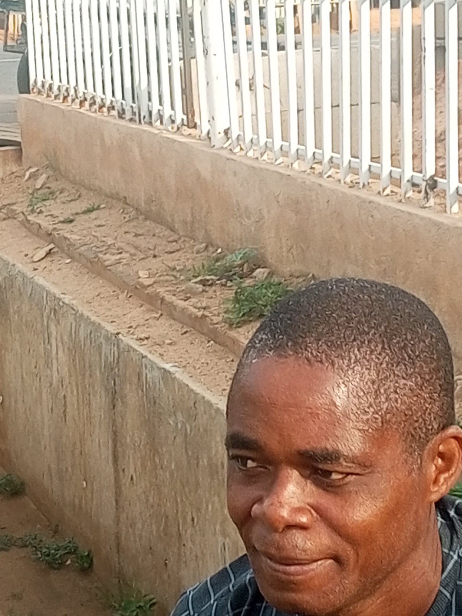 A medium shot captures a man's face and upper torso, with a blurred background of a white metal fence and a concrete structure. The man, with dark skin and short, textured hair, is looking to his left. His expression is neutral, with his mouth slightly pursed. He is wearing a dark plaid shirt. The background consists of vertical white bars of a fence, positioned behind a textured, beige concrete wall. Some patches of dry vegetation are visible on the ground and along the wall. The lighting suggests it is daytime.
