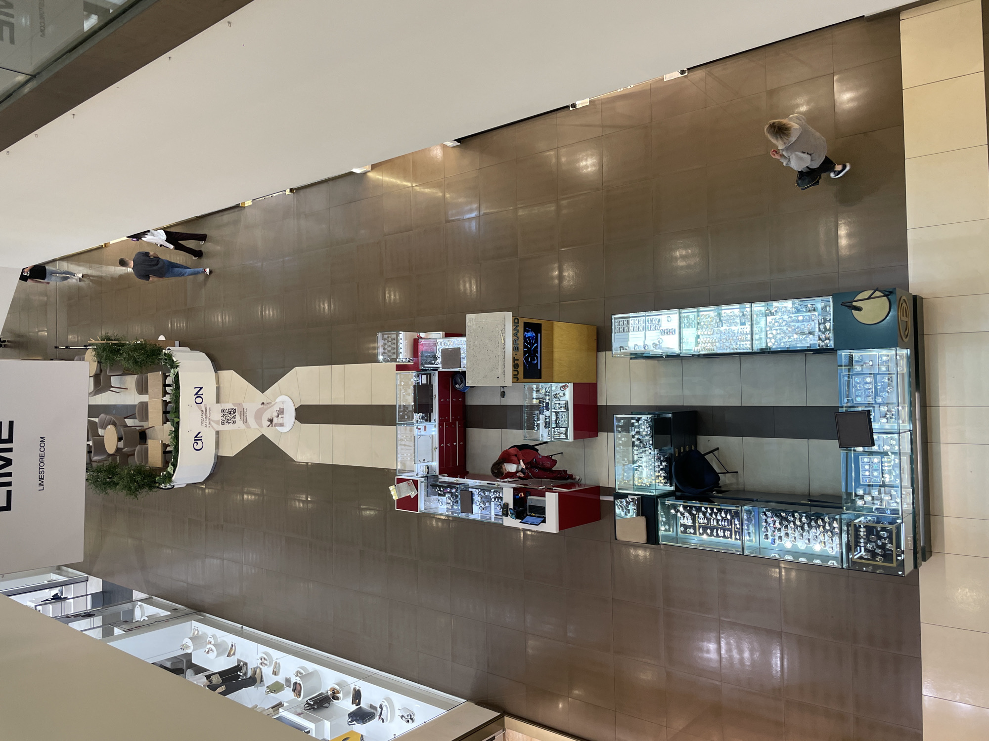 The image depicts an aerial view of a shopping mall interior. A retail space called "LIME" is visible on the left, with shoe displays and a lounge area. In the center, there's a promotional area for "CINQSON" with displays and seating. Further down the mall, there are jewelry kiosks showcasing watches and other accessories. People are seen walking through the mall. The scene is indoors with artificial lighting, and the floors are tiled. The location appears to be Saint Petersburg, Russia, given the Cyrillic lettering on the CINQSON display.