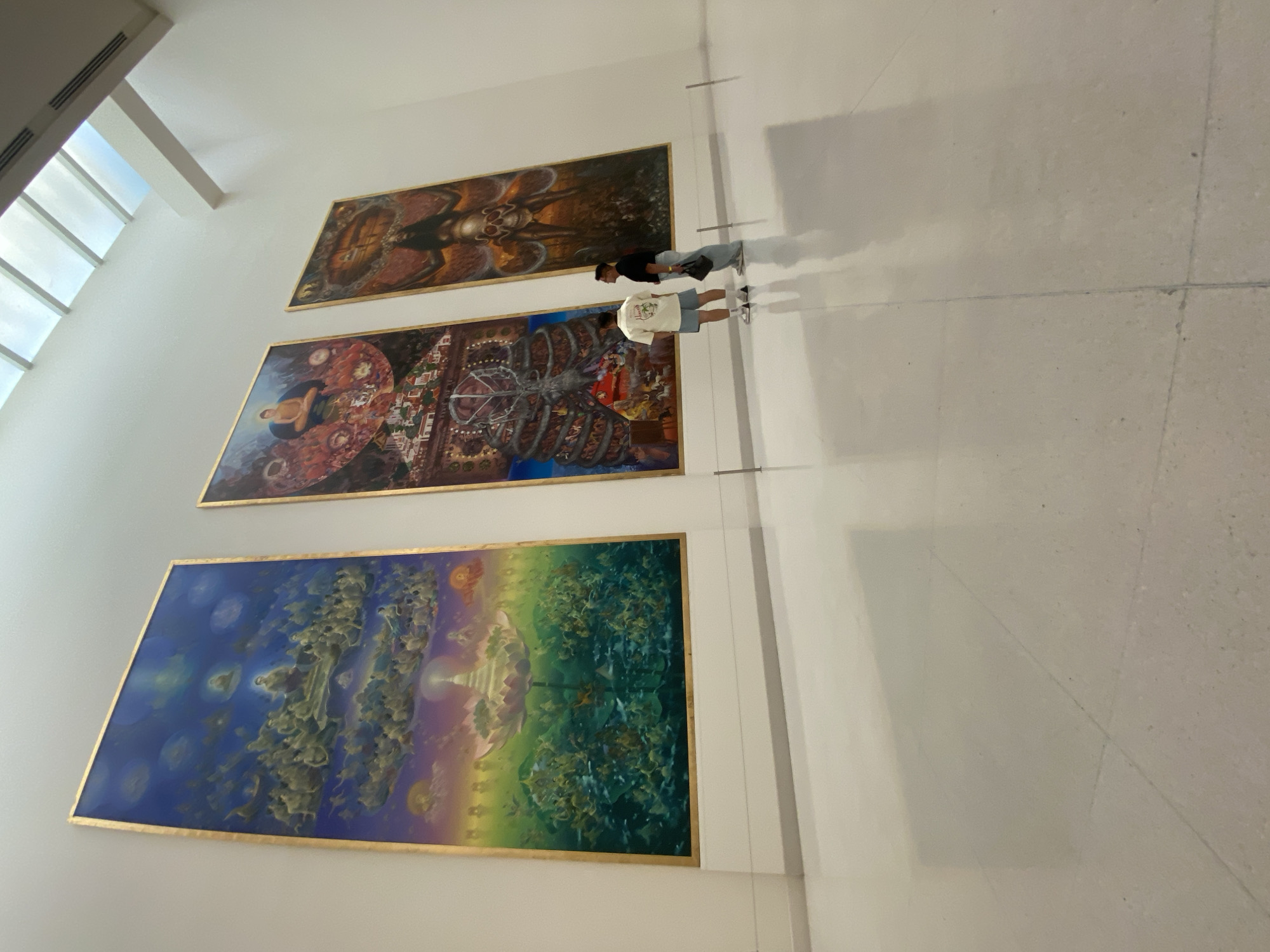 The photograph shows three large paintings hanging on a white wall in what appears to be an art gallery or museum. Two individuals are standing near the paintings. The paintings are of varying sizes and styles; the bottom painting is the largest and features blue, green, and yellow colors, with complex designs. The middle painting has a reddish-brown color scheme and intricate patterns, while the top painting features dark brown and black tones. The environment appears to be well-lit and modern, with natural light coming in from a nearby window.
Based on the art style of the paintings and the gallery architecture, the location is likely the Museum of Contemporary Art (MOCA) in Bangkok, Thailand. The art style strongly resembles that of Thai artist Thawan Duchanee.