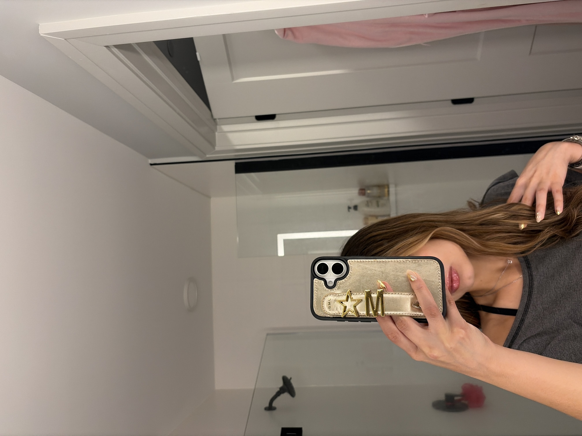 A woman is taking a selfie in a bathroom, likely in a residence in Austin, Texas. The bathroom features a white wall, a mirror reflecting the woman and the bathroom fixtures, including a faucet and potentially a shower head. The woman is holding a smartphone with a distinctive case adorned with a star and the letter "M," partially obscuring her face. Her hair is light brown with highlights. She is wearing a gray top and a necklace. The reflection in the mirror reveals additional bathroom elements, such as toiletries or decorative items on a shelf. Above the mirror, there appears to be a white cabinet or door frame. A pink item is visible on the top edge of the frame. The image is taken at an angle, giving a view of the ceiling.