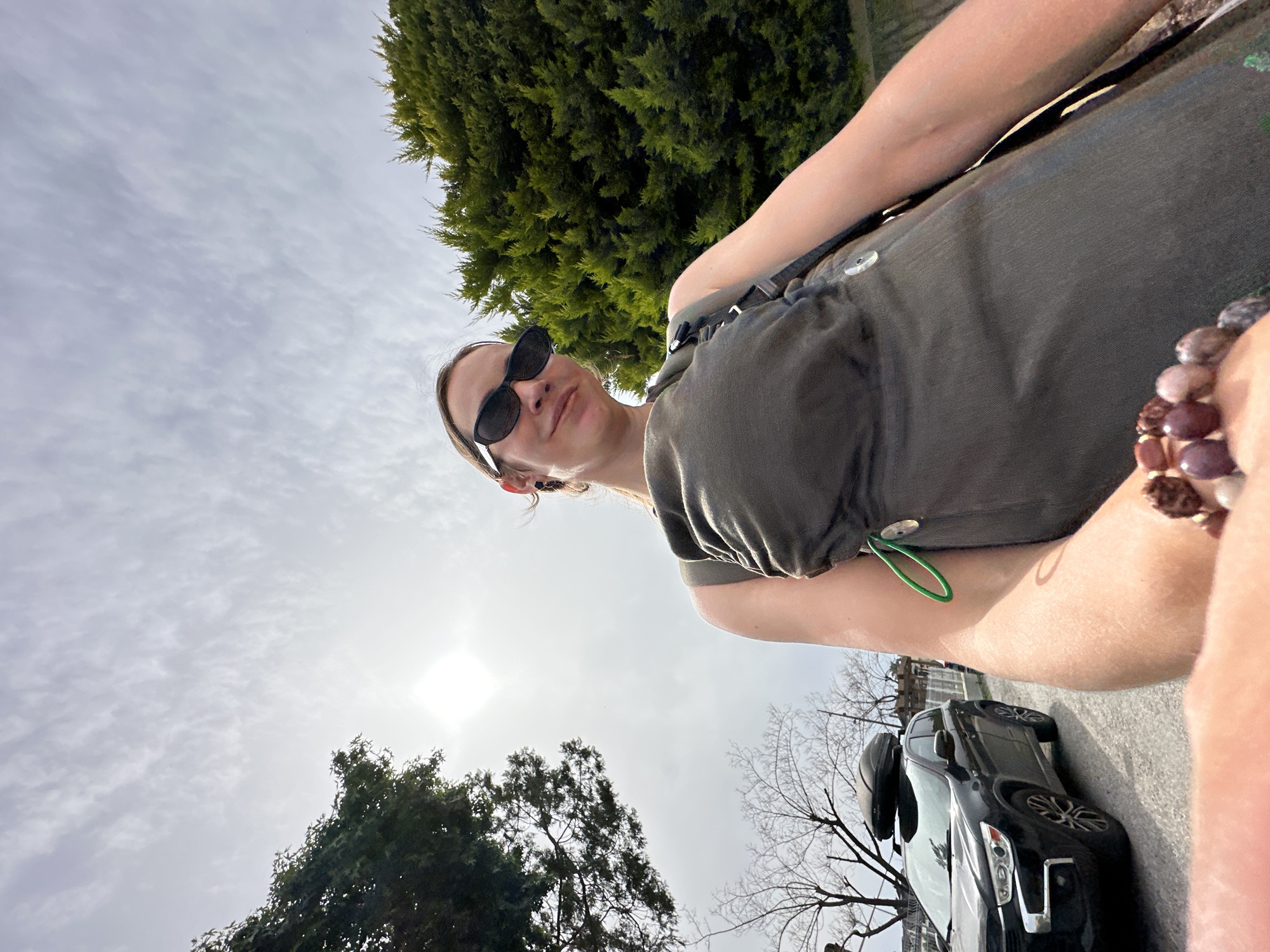 A photograph taken outdoors shows a person wearing sunglasses and an olive-green, button-down shirt. The person is positioned diagonally, with the sky and the sun visible in the upper left portion of the frame. There is a green tree behind the person, and a black car parked on a paved road in the lower right portion of the frame. The photograph appears to be taken from a low angle. The location in Fethiye, Turkey cannot be determined from the image.