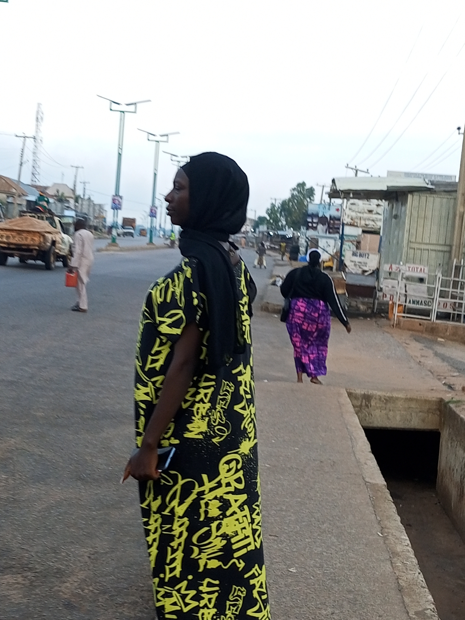 A medium shot shows a woman wearing a black hijab and a yellow graffiti-patterned dress standing on a concrete sidewalk in Gombe, Nigeria. Her face is partially visible as she looks to her left, and her right hand holds a dark-colored smartphone. In the background, a street with traffic is visible, including a sand-filled truck, a white pickup truck, and several other vehicles and people. Lampposts with solar panels line the street, and buildings with corrugated metal roofs are on the right side of the image. A dark, open drainage ditch is located on the lower right of the frame. Another woman, wearing a black top and a purple and pink patterned skirt, walks away from the camera on the sidewalk. The sky is overcast, suggesting it is either early morning or late afternoon.