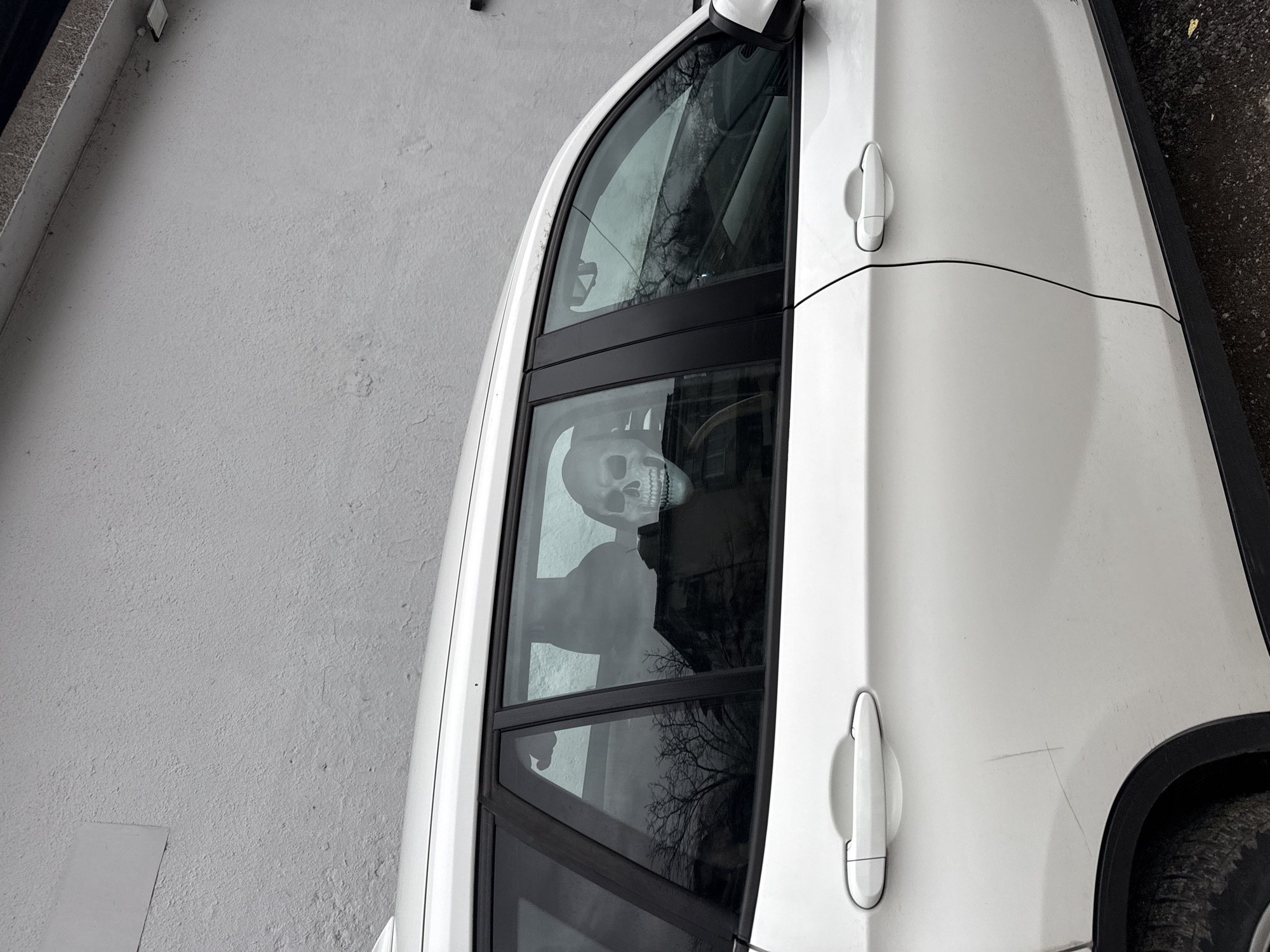 A white car, possibly a crossover or SUV, is parked next to a light gray or white concrete wall. The car faces toward the right side of the image. Inside the car, in the driver's seat, there is a white skull. The scene is likely in an urban setting, possibly in Sofia, Bulgaria, based on general architectural styles. The reflection in the car window shows what appears to be a building and bare tree branches.