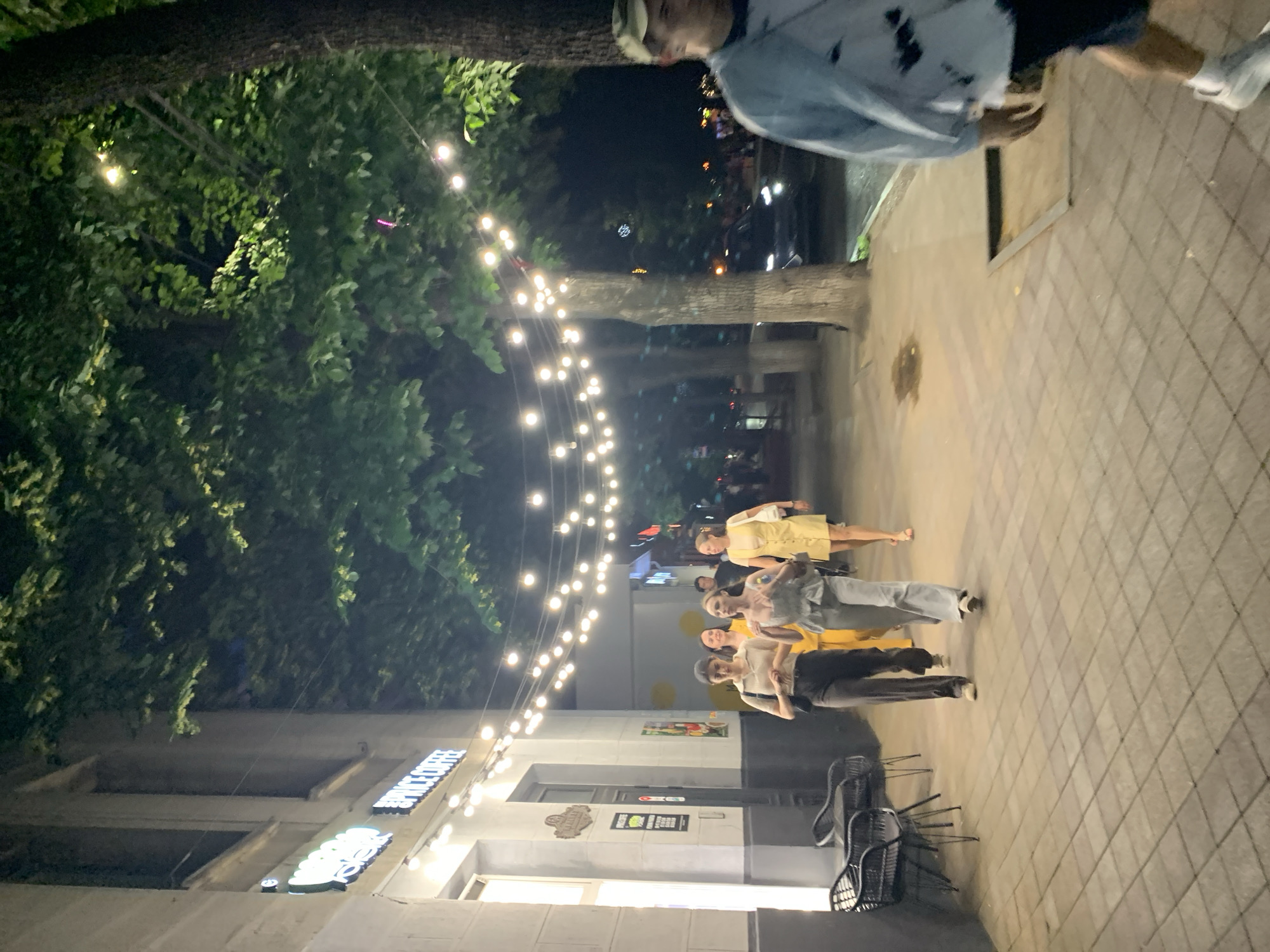 The image depicts a street scene at night in an urban setting, with the location identified as Unknown, Unknown. A building with a sign that reads "SPICE COFFEE" is visible on the left, illuminated by string lights that stretch across the street, suspended from trees. Several people are walking on the sidewalk in front of the building. On the right side of the image, a man in a light-colored jacket and a baseball cap is partially visible, walking away from the camera. The sidewalk is paved with square tiles. The environment is characterized by the presence of trees with dense foliage, street lights, and parked cars in the background. The overall atmosphere suggests a lively evening in a city.