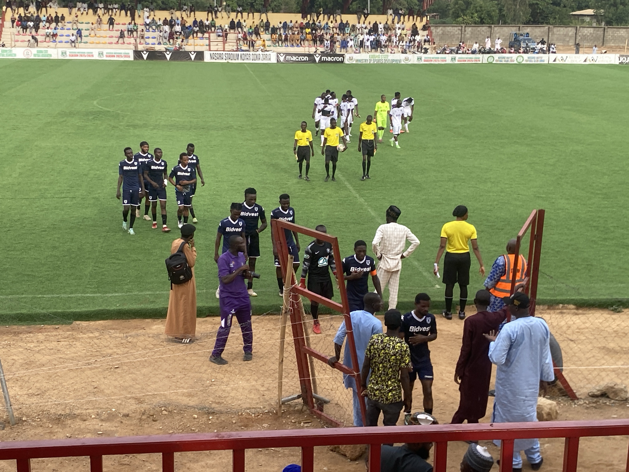The image depicts a soccer match at Nasara Stadium Kofar Doka Zaria, in what appears to be Tudun Wada, Nigeria. The field is green and teams dressed in dark blue and white are present, along with referees in yellow. Spectators are in the stands. Individuals are seen near a fenced area in the foreground. Branding for "Bidvest" is visible on the uniforms. Signage behind the field indicates the season is 2025/2026.