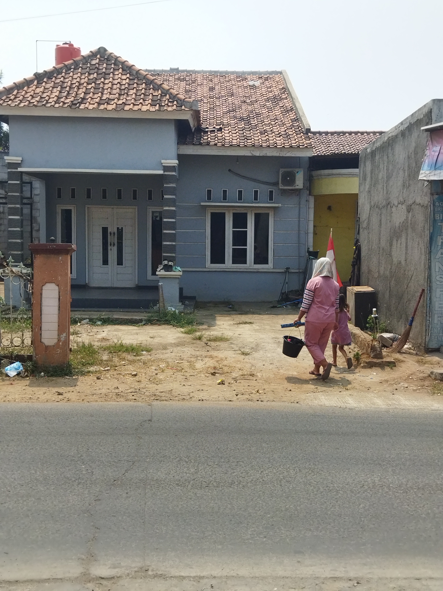 The image depicts a residential scene in Suka Sari, Indonesia. In the foreground, a paved road runs horizontally across the frame. Beyond the road, a dirt path leads to a modest light blue house with a terracotta tile roof. A red water tank is visible on the roof. The house has white double doors and several windows. To the right of the house is a yellow building and a tall, gray concrete wall.
Two individuals, an adult and a child, are walking away from the house towards the right side of the image. The adult, wearing a pink headscarf and matching pink outfit, carries a black bucket in their right hand. The child, wearing a light purple dress, walks slightly ahead. A broom leans against the gray wall. The ground around the house and path is a mix of dirt, sparse vegetation, and scattered debris. The sky is clear and bright.