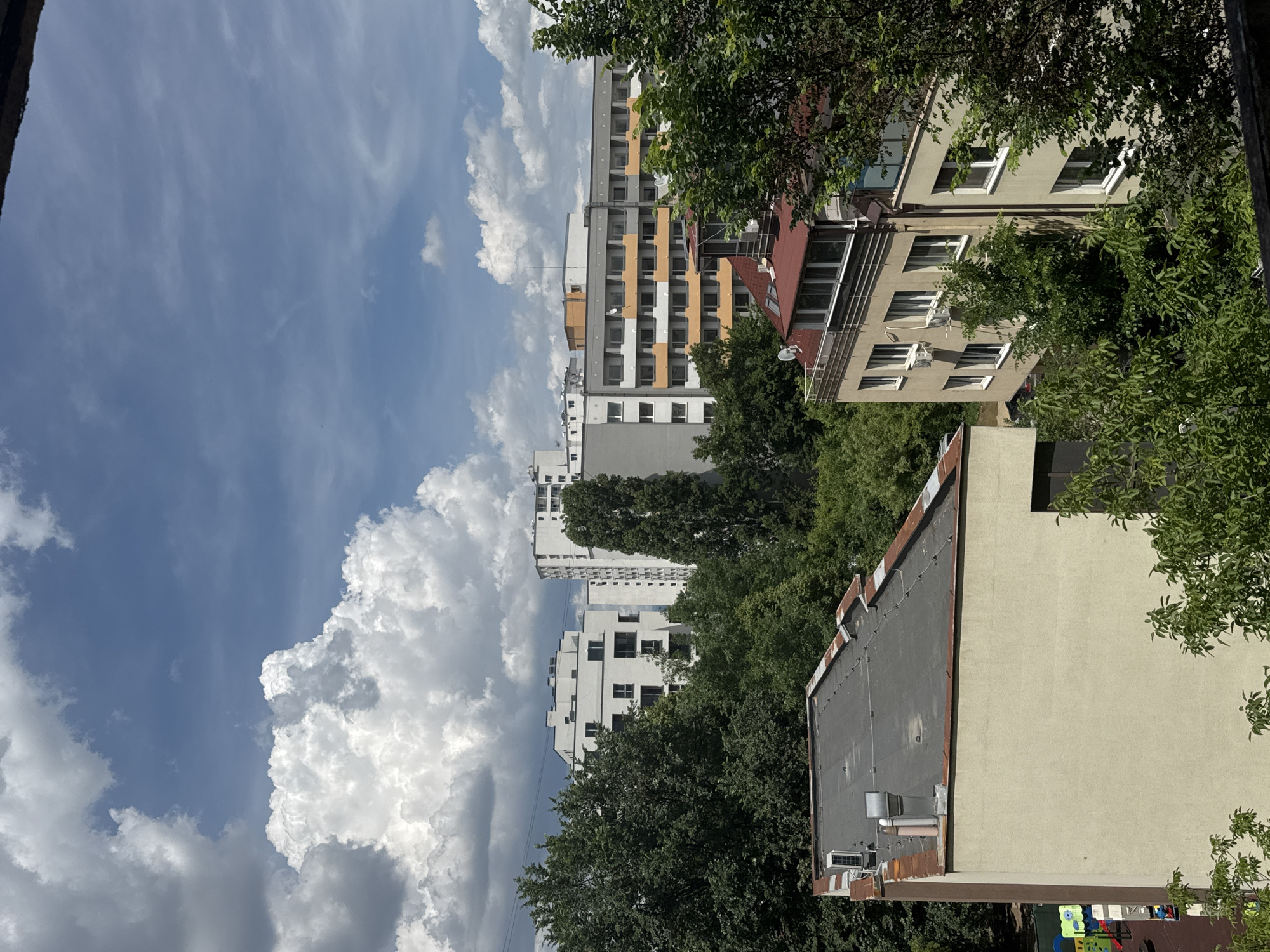 The image captures a view of a residential area in Sofia, Bulgaria, under a partially cloudy sky. In the foreground, the flat roof of a light-colored building is visible, equipped with an air conditioning unit and a ventilation pipe. Adjacent to this building and slightly further back is another building with a distinctive red-tiled roof, featuring multiple windows and a balcony.
The middle ground is dominated by dense green trees, interspersed with several multi-story apartment buildings. One prominent building is gray with orange balconies, showing many windows. To the left, there's a white building with a modern design, also featuring numerous windows and a flat roof. Further in the distance, more white apartment buildings can be seen. The sky above is a mix of bright blue and large, fluffy white clouds, characteristic of a sunny day. The overall impression is a typical urban landscape with a blend of residential architecture and abundant greenery.