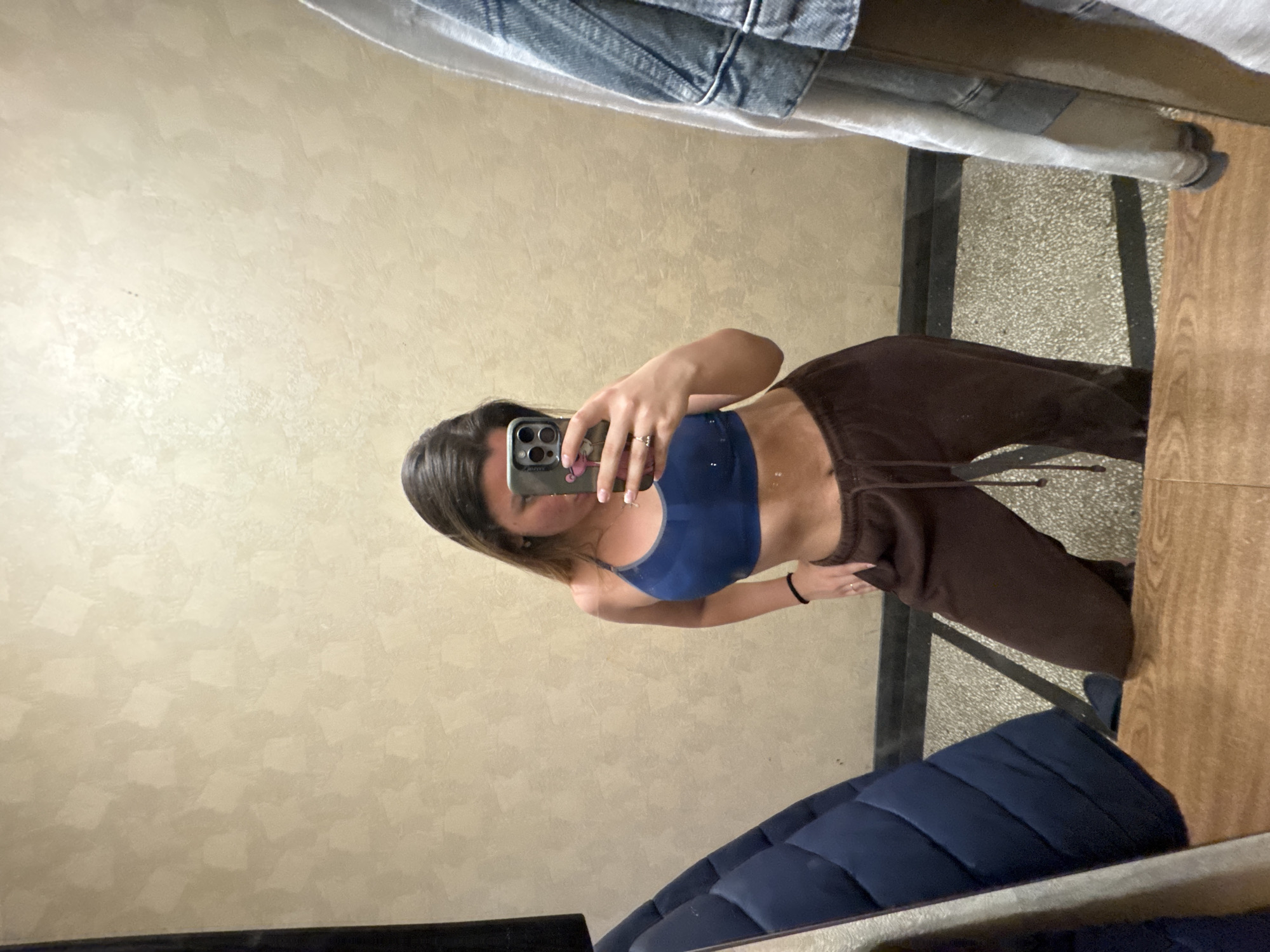 A mirror selfie shows a person with long brown hair and a fair complexion wearing a navy blue sports bra and brown sweatpants. The person is holding an iPhone with a phone case that appears to have images on it. The person is wearing a ring on their left hand and a black bracelet on their right wrist. The reflection shows part of a tan wall with a textured finish, a blue quilted object resembling a cushion or possibly part of a coat, and part of what seems to be a wooden table or shelf. Also in the reflection are articles of clothing and what appears to be light-colored flooring. There is insufficient information in the image to determine the location as Sofia, Bulgaria.