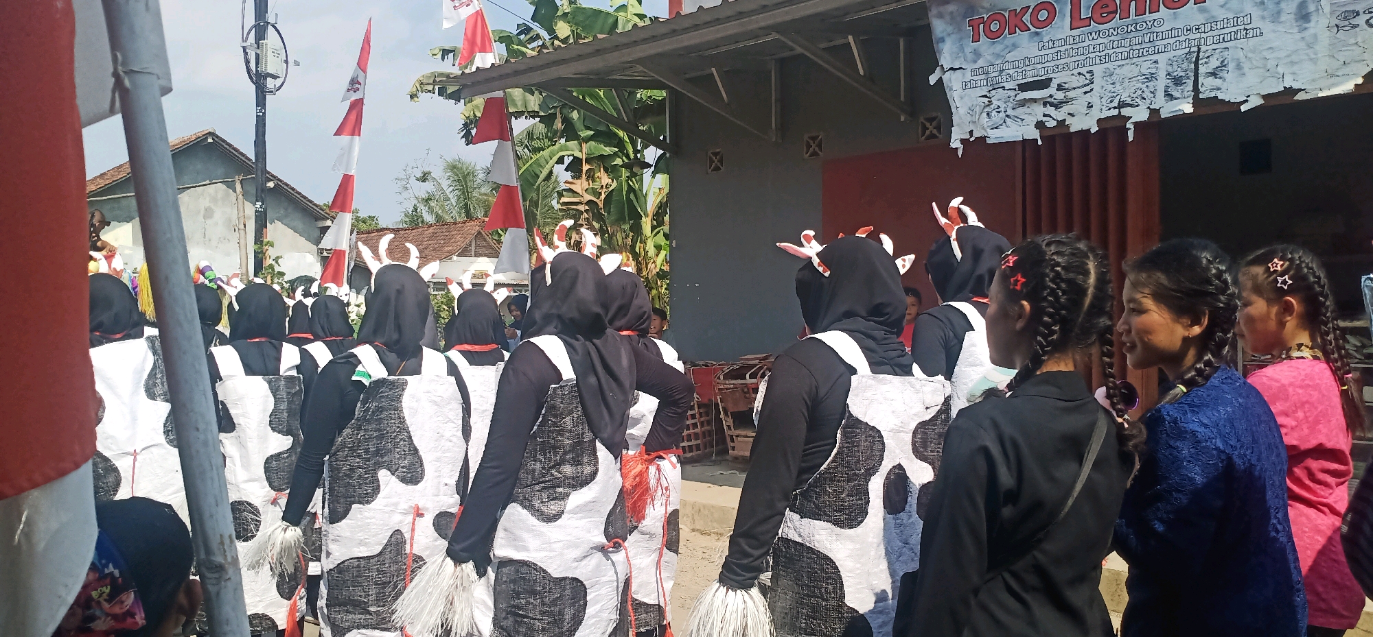 The image captures a street scene in Kincang, Indonesia, likely during a parade or cultural event. In the foreground, a group of individuals, mostly young women, are dressed in black outfits and appear to be costumed as cows. They wear black head coverings with white and red cow-like horns. Their costumes feature a black and white spotted pattern, resembling cowhide. Some of them have white fringes hanging from their waists. Behind them, several girls with braided hair, adorned with star-shaped clips, are visible. One girl wears a blue top, another a pink top, and one a black jacket. In the background, a building with a corrugated metal roof is adorned with Indonesian flags. A banner with Indonesian text, including "TOKO LENO," is also visible. The environment suggests a rural or semi-urban setting with houses and trees. The overall atmosphere appears festive and communal.